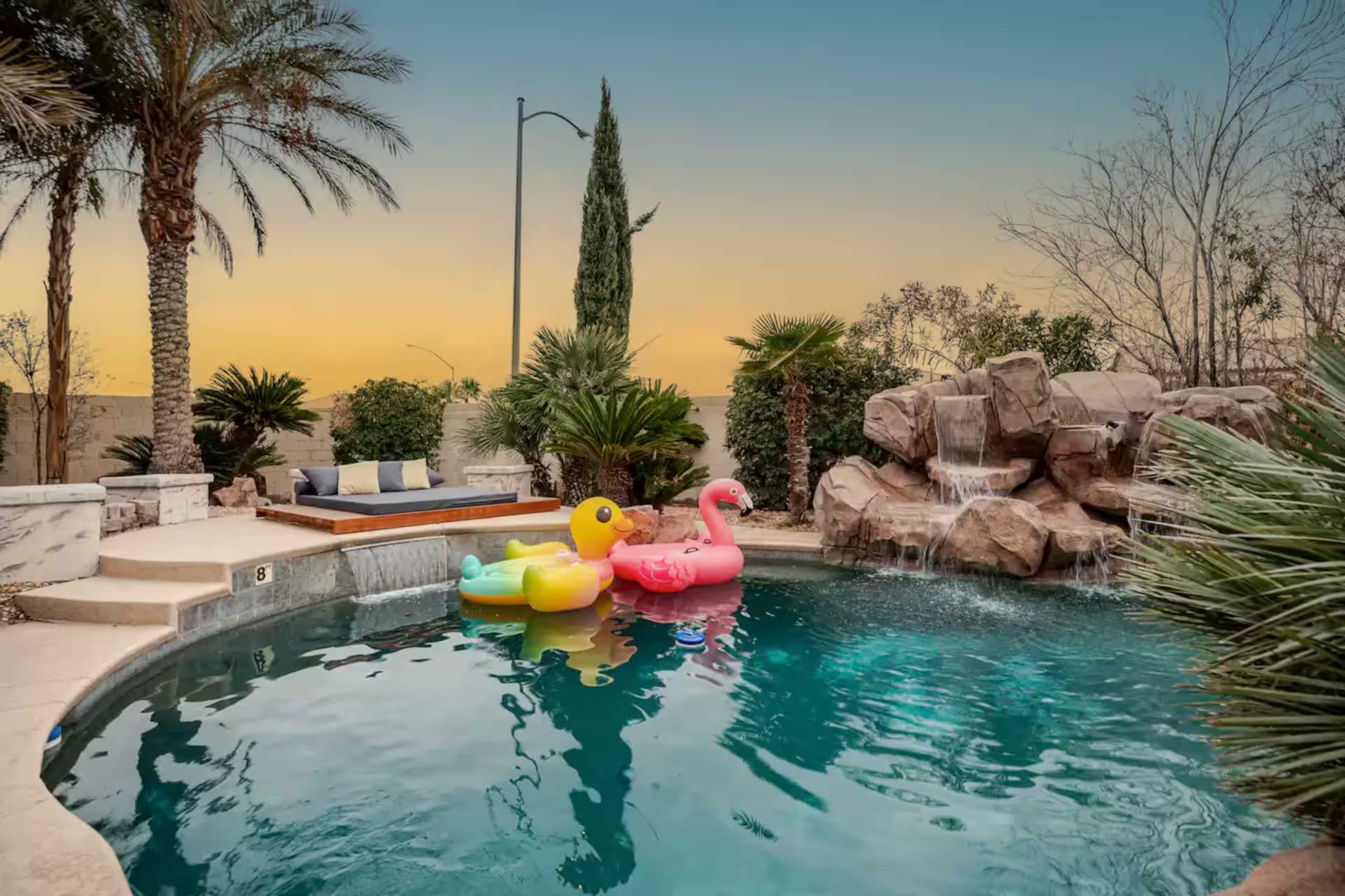 The scene features a swimming pool with colorful floaties, surrounded by palm trees and a rock waterfall, under a sunset sky.