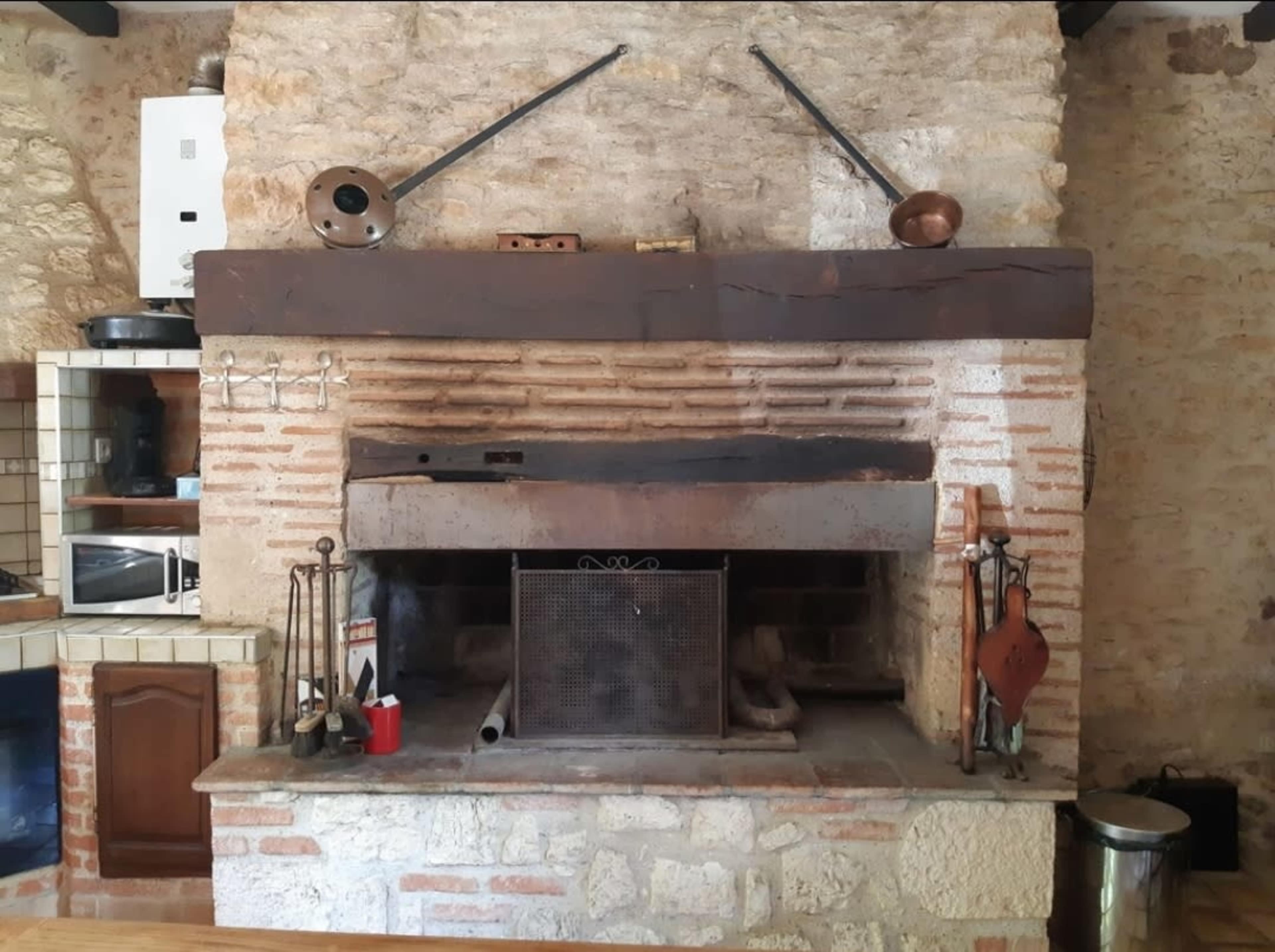 The image shows a rustic stone-built fireplace with a metal grate and various cooking tools arranged around it in a kitchen setting.