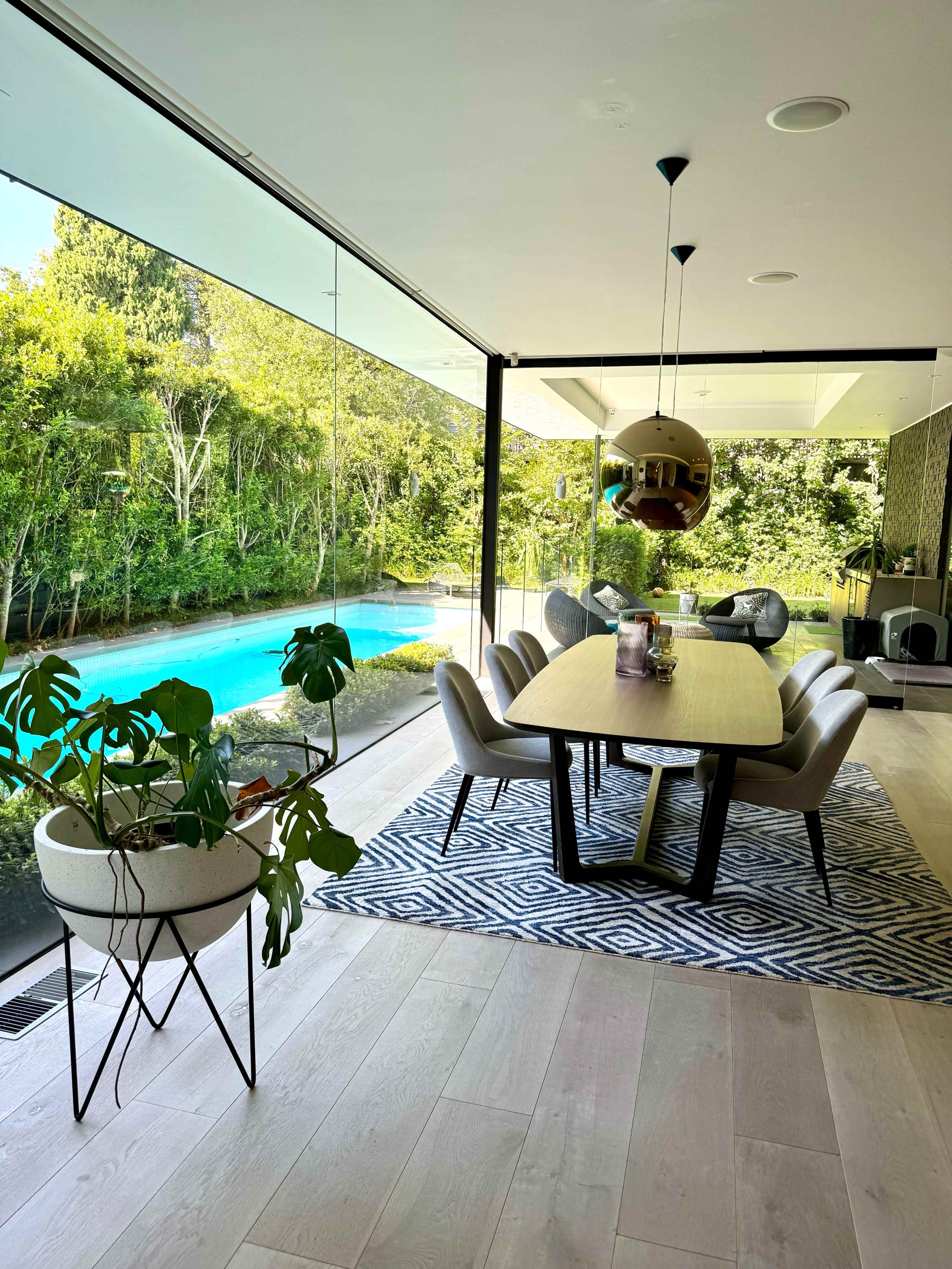 A modern dining area features a large table surrounded by chairs, with a view of a blue swimming pool and greenery through glass walls.