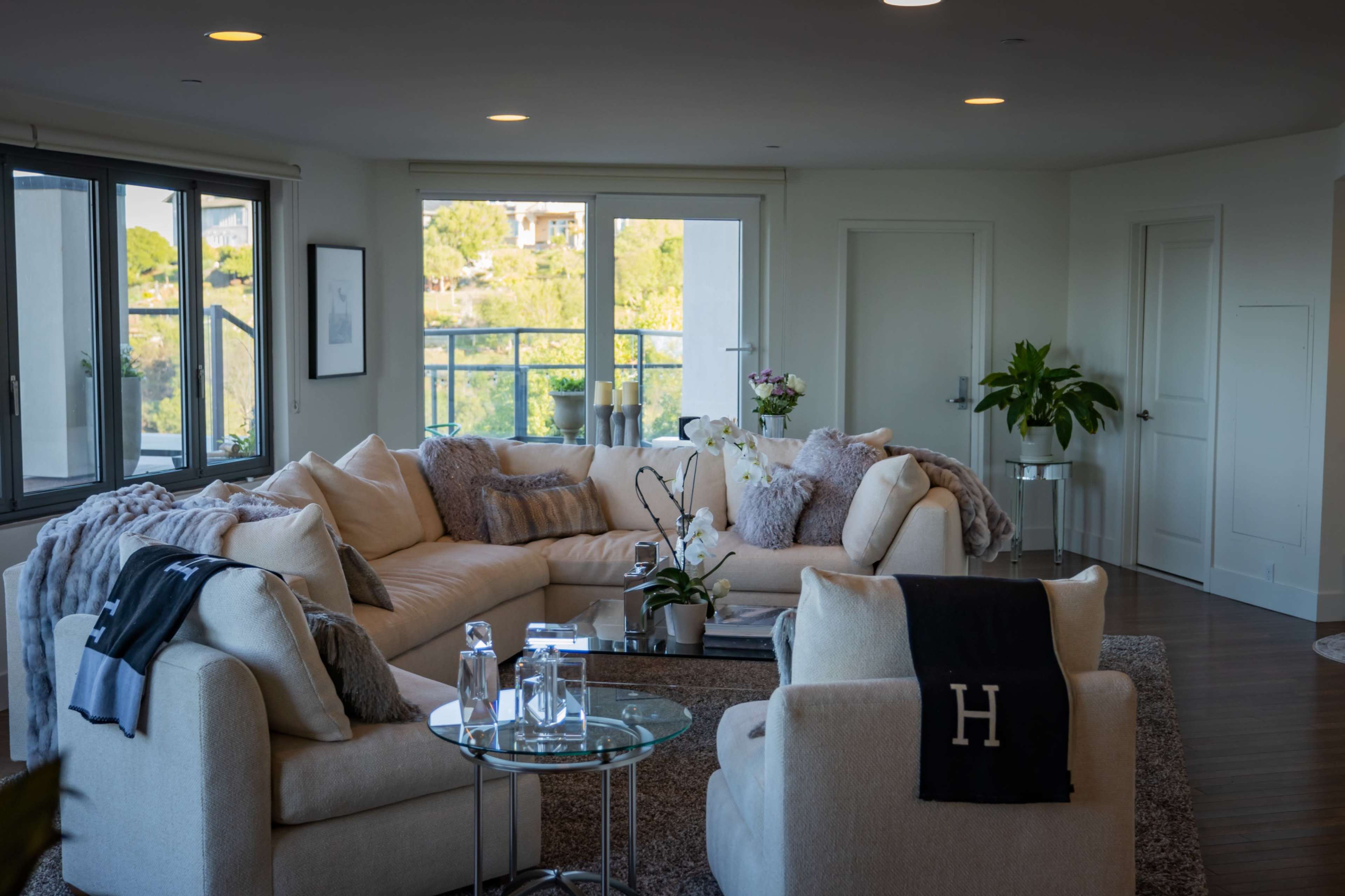 A spacious living room features a large, L-shaped sectional sofa with light-colored cushions and a glass coffee table, set against large windows overlooking a green landscape.