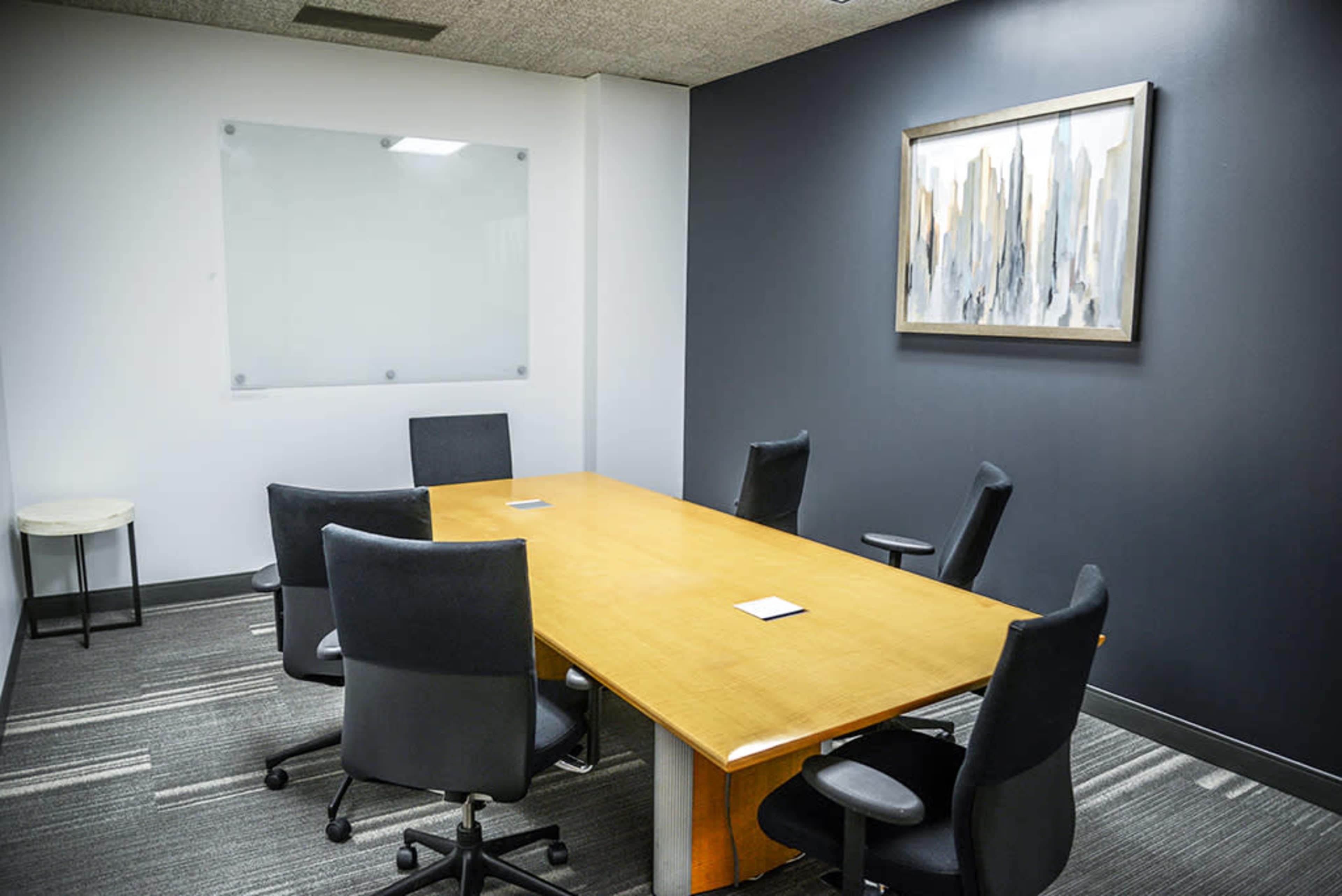 A conference room features a large wooden table surrounded by six black office chairs, with a whiteboard and a framed abstract painting on the walls.