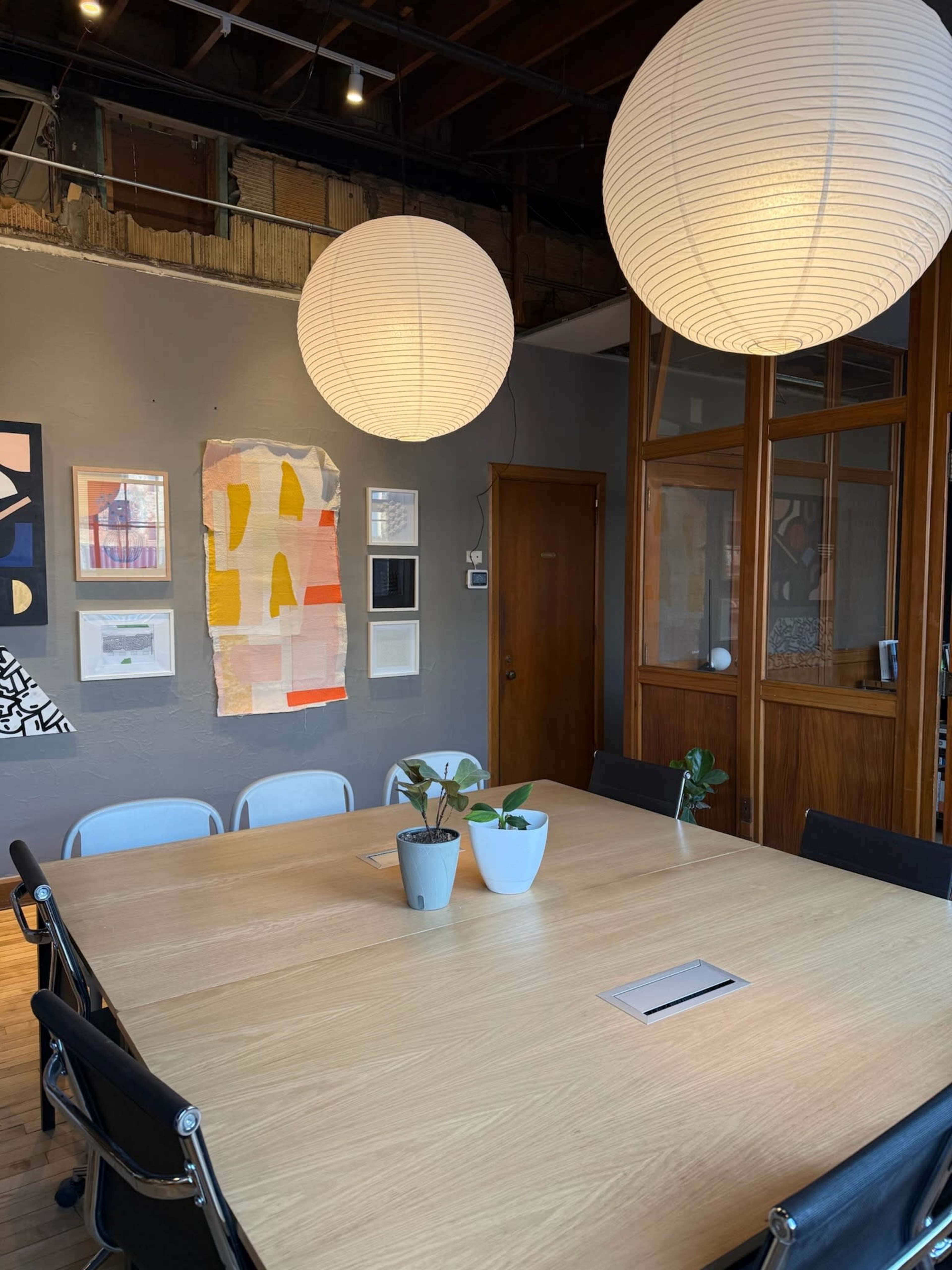 The image shows a modern conference room with a large wooden table, two hanging light fixtures, and artwork displayed on the walls.