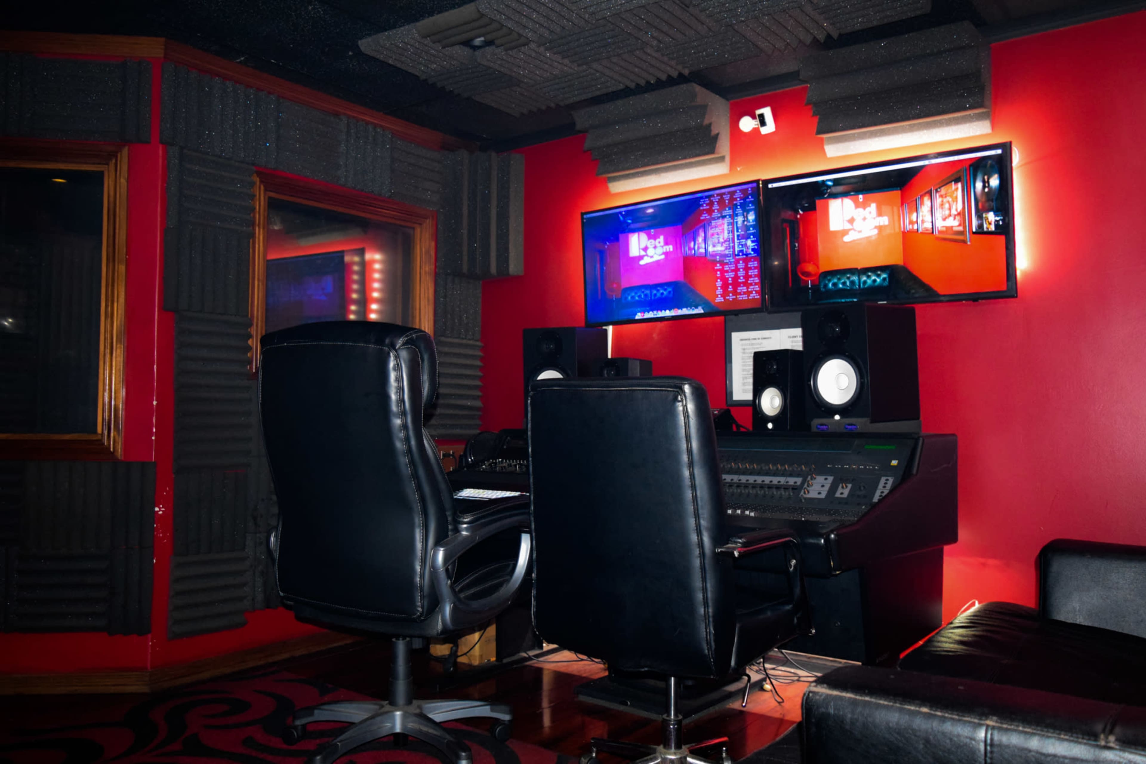 The image shows a sound recording studio equipped with a mixing console, two monitors, and black leather seating against a red wall.