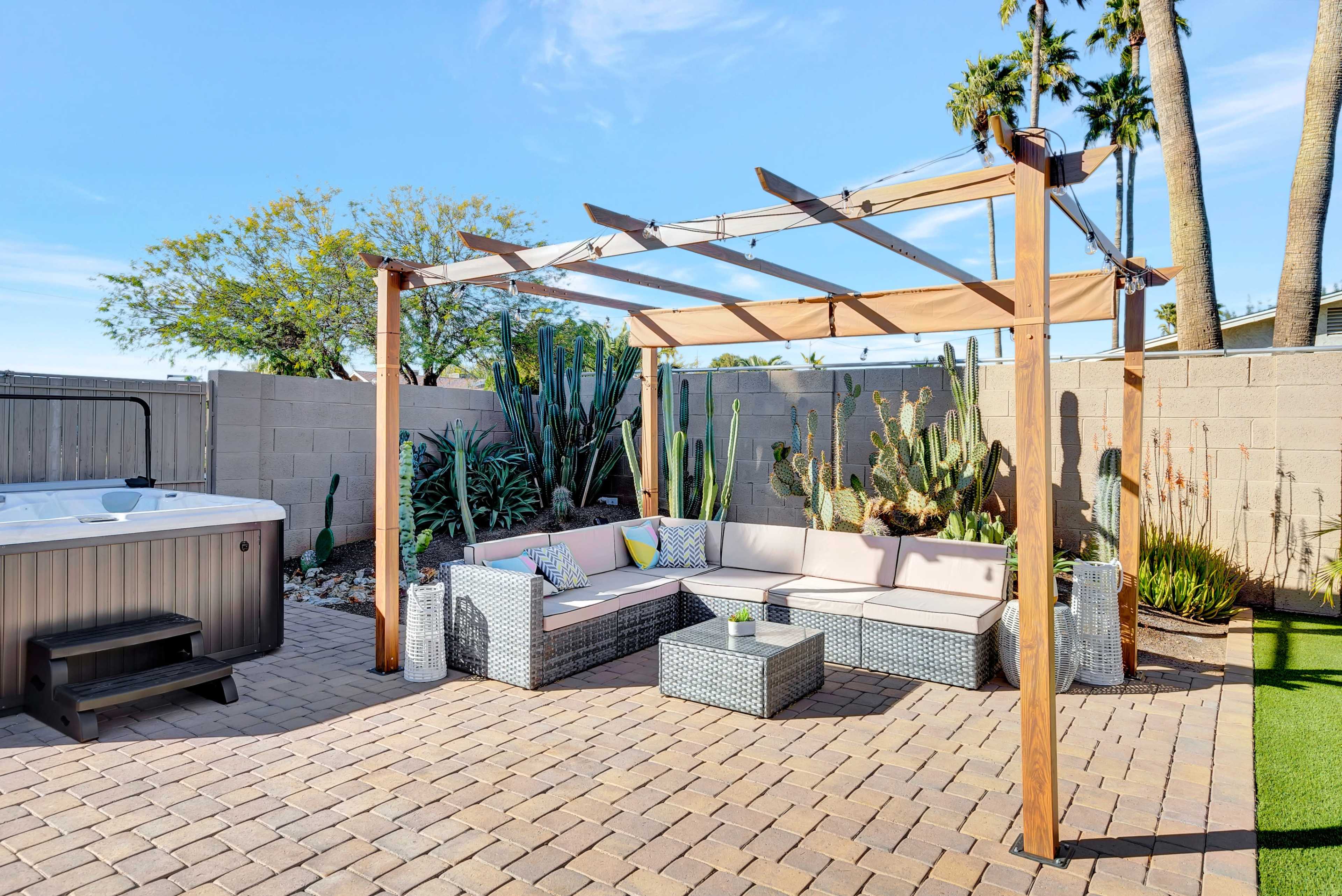 A covered outdoor seating area with a sectional couch and coffee table is located beside a hot tub, surrounded by cacti and palm trees.