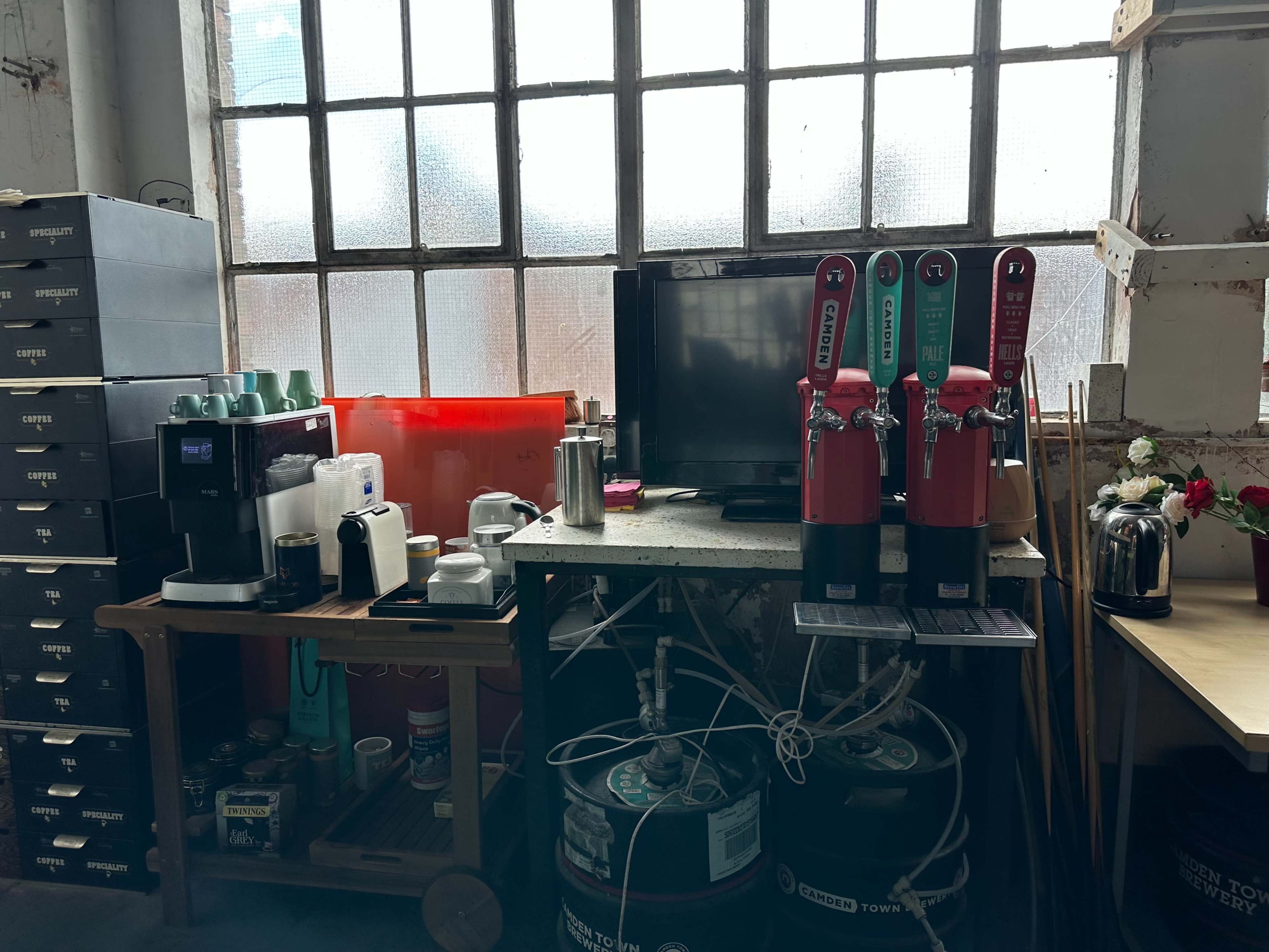 A break area with a countertop featuring coffee machines, a television, and beverage dispensers, against a backdrop of large, industrial windows.