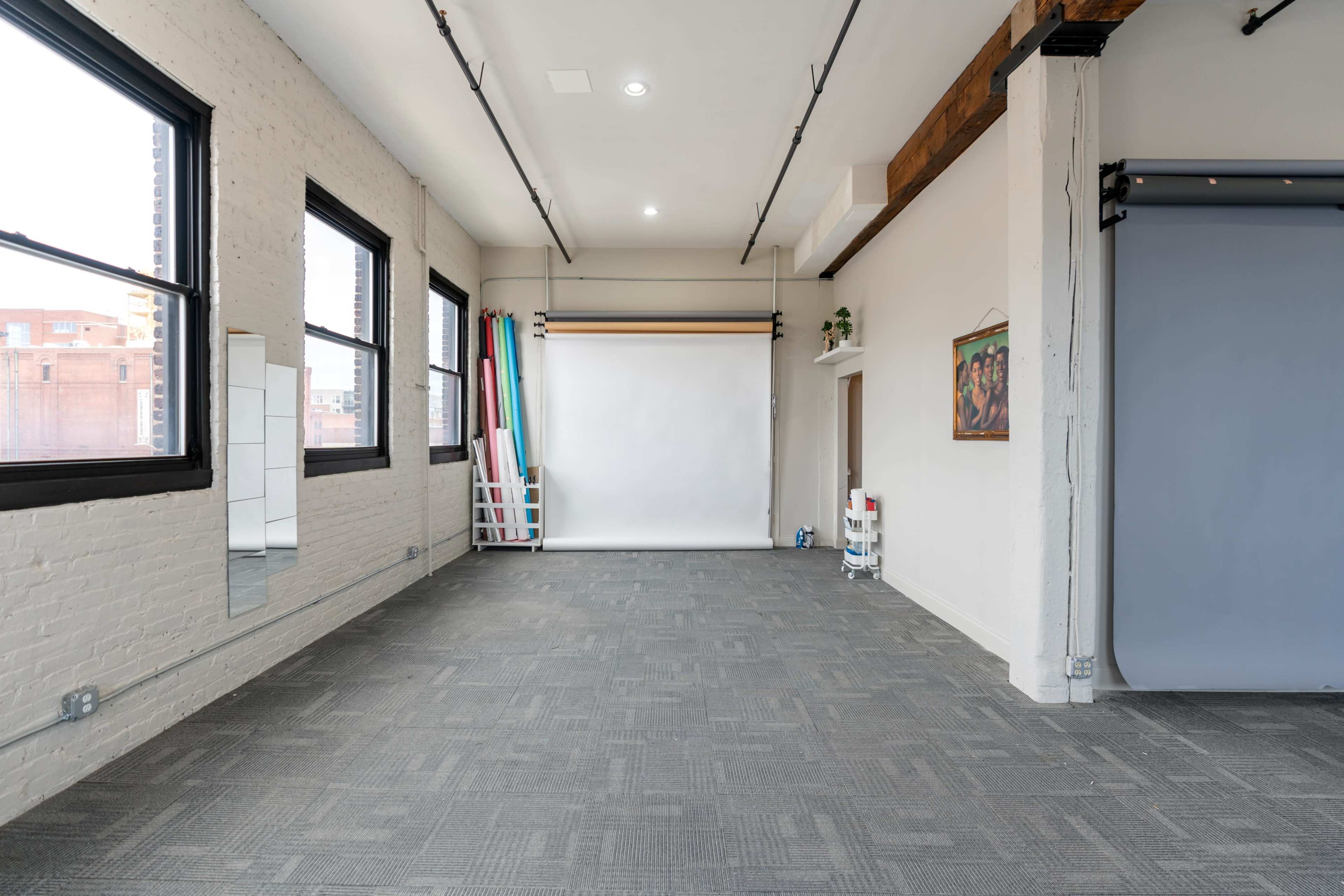 A spacious, empty studio with large windows, a white backdrop, and rolled backdrops in various colors against one wall.