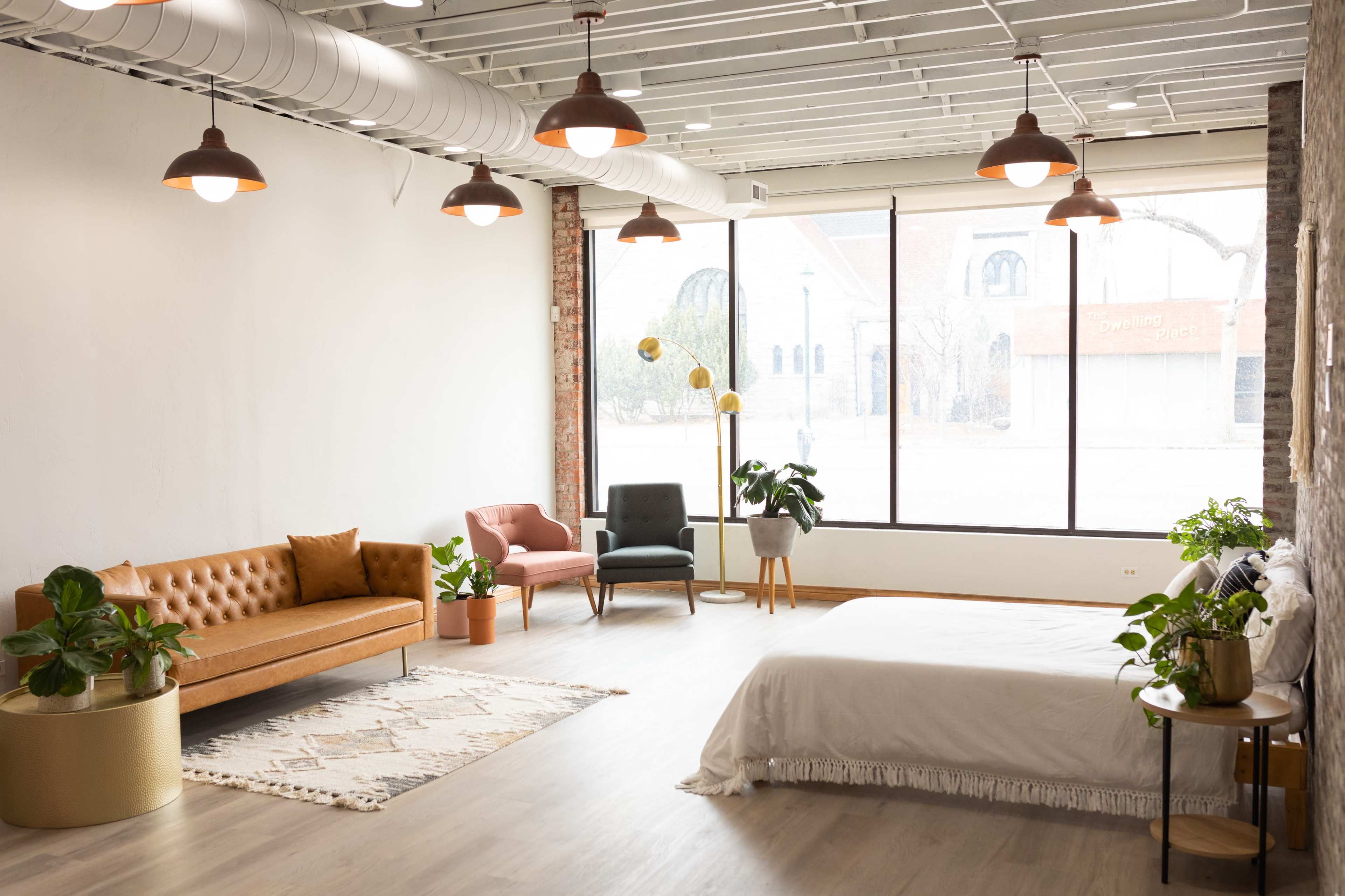A spacious, modern room featuring a bed, a sofa, two armchairs, and various indoor plants, with large windows allowing natural light to flood the space.