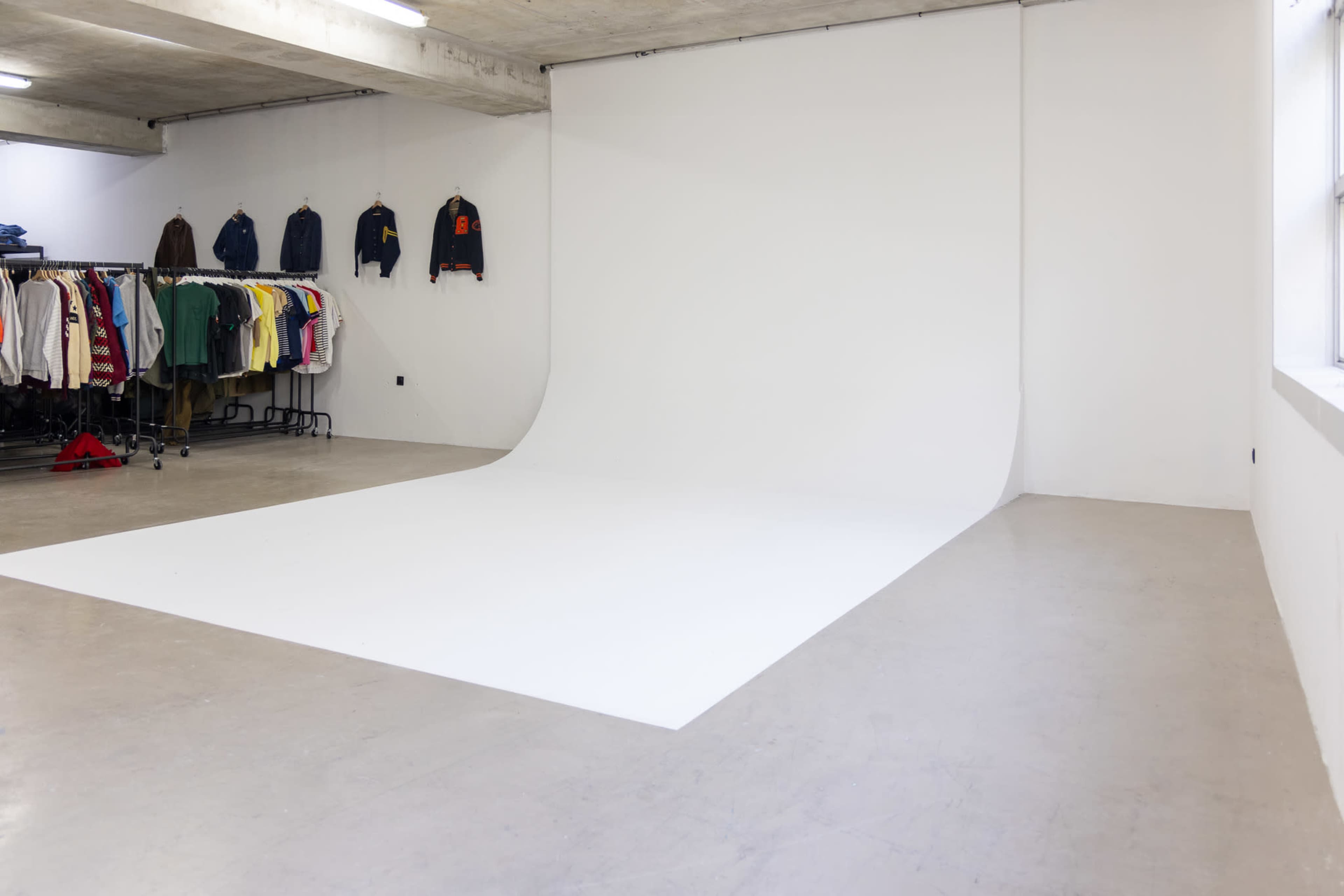 The image shows a spacious, minimalist studio with a white backdrop and a rack of colorful clothing hanging on a metal frame.