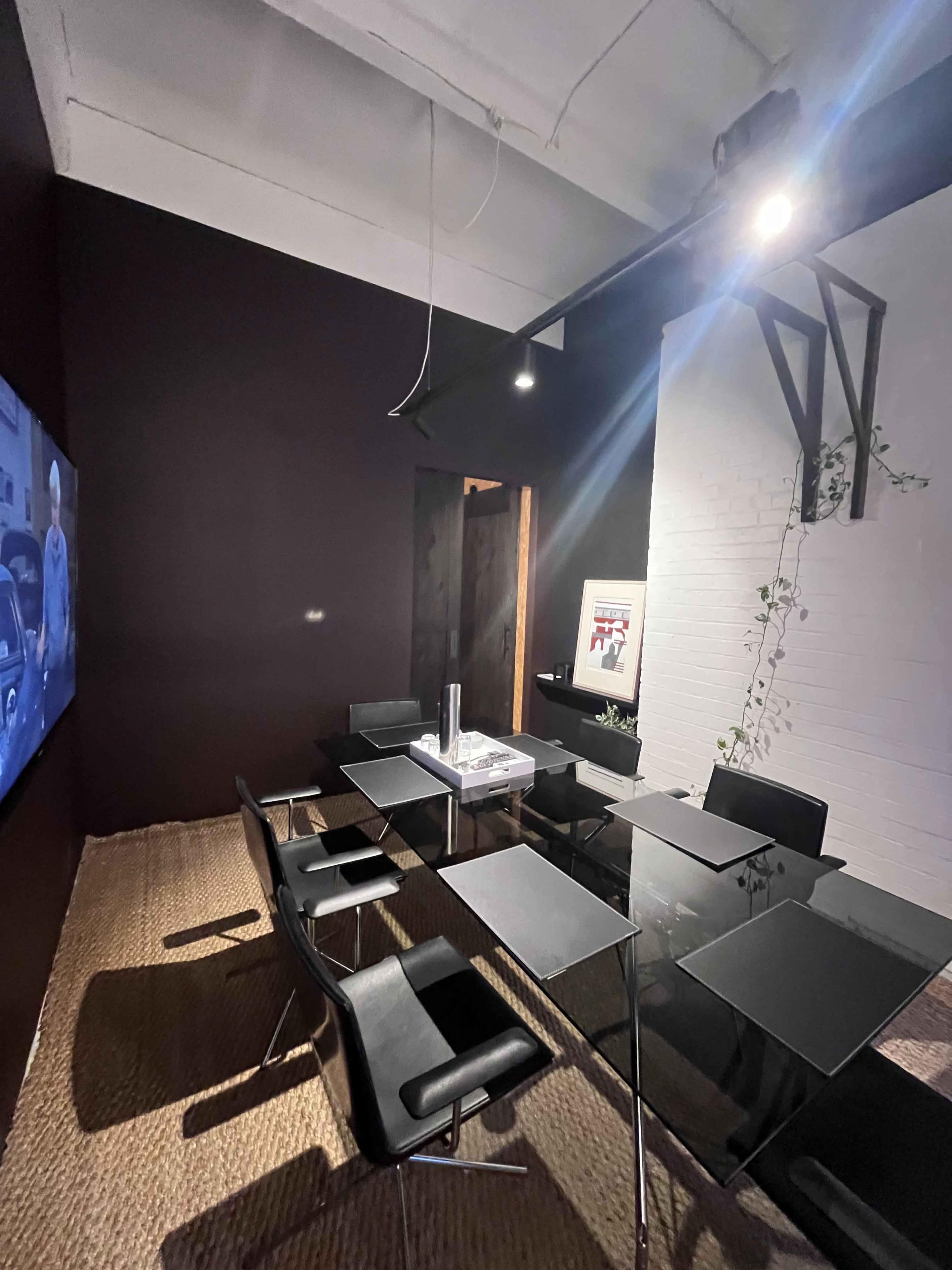 The image shows a modern conference room with a glass table surrounded by black chairs, a wall-mounted screen, and minimalist decor.