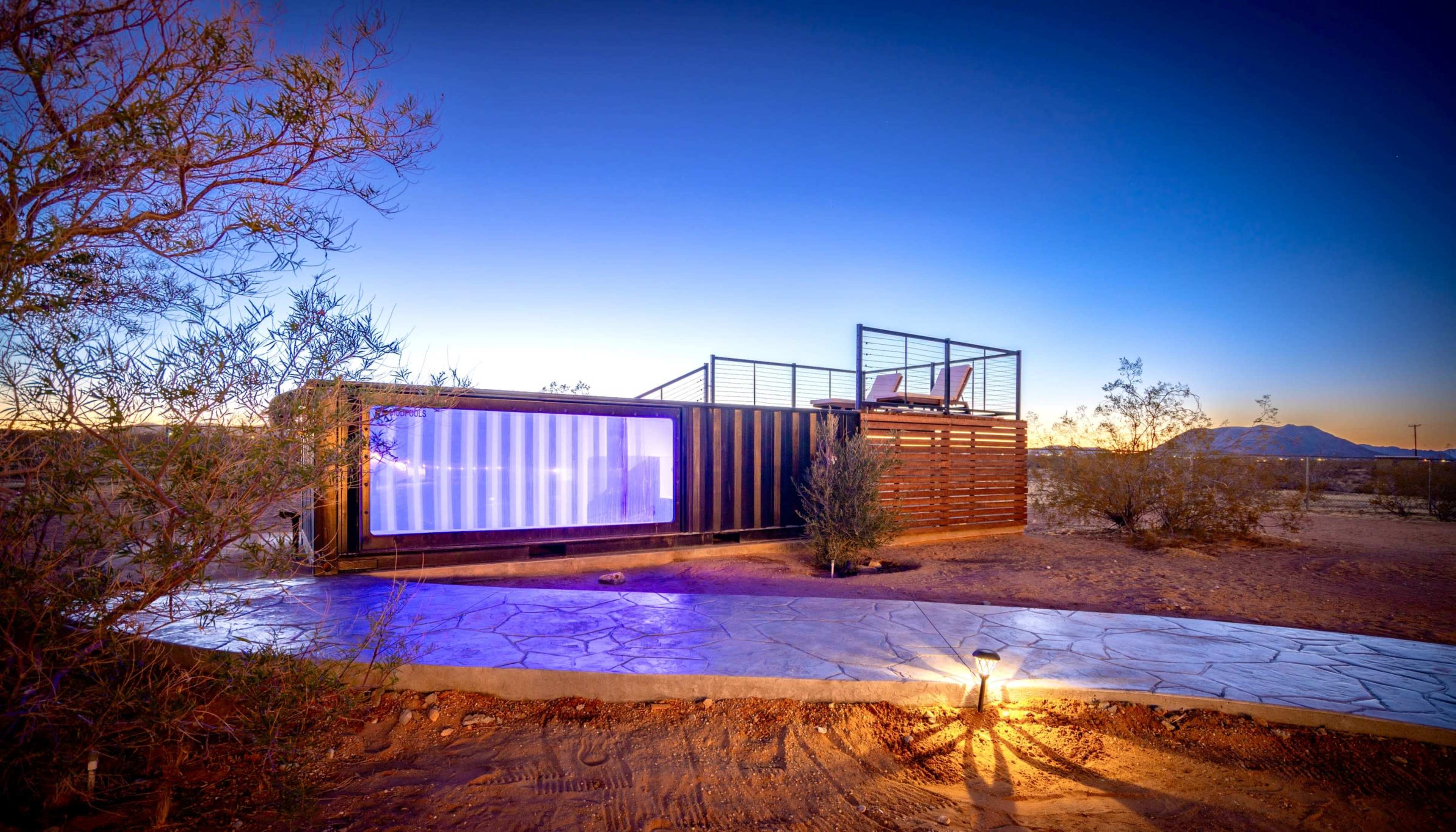 A geometric structure made from shipping containers stands in a desert landscape under a twilight sky, with a concrete pathway and low vegetation nearby.