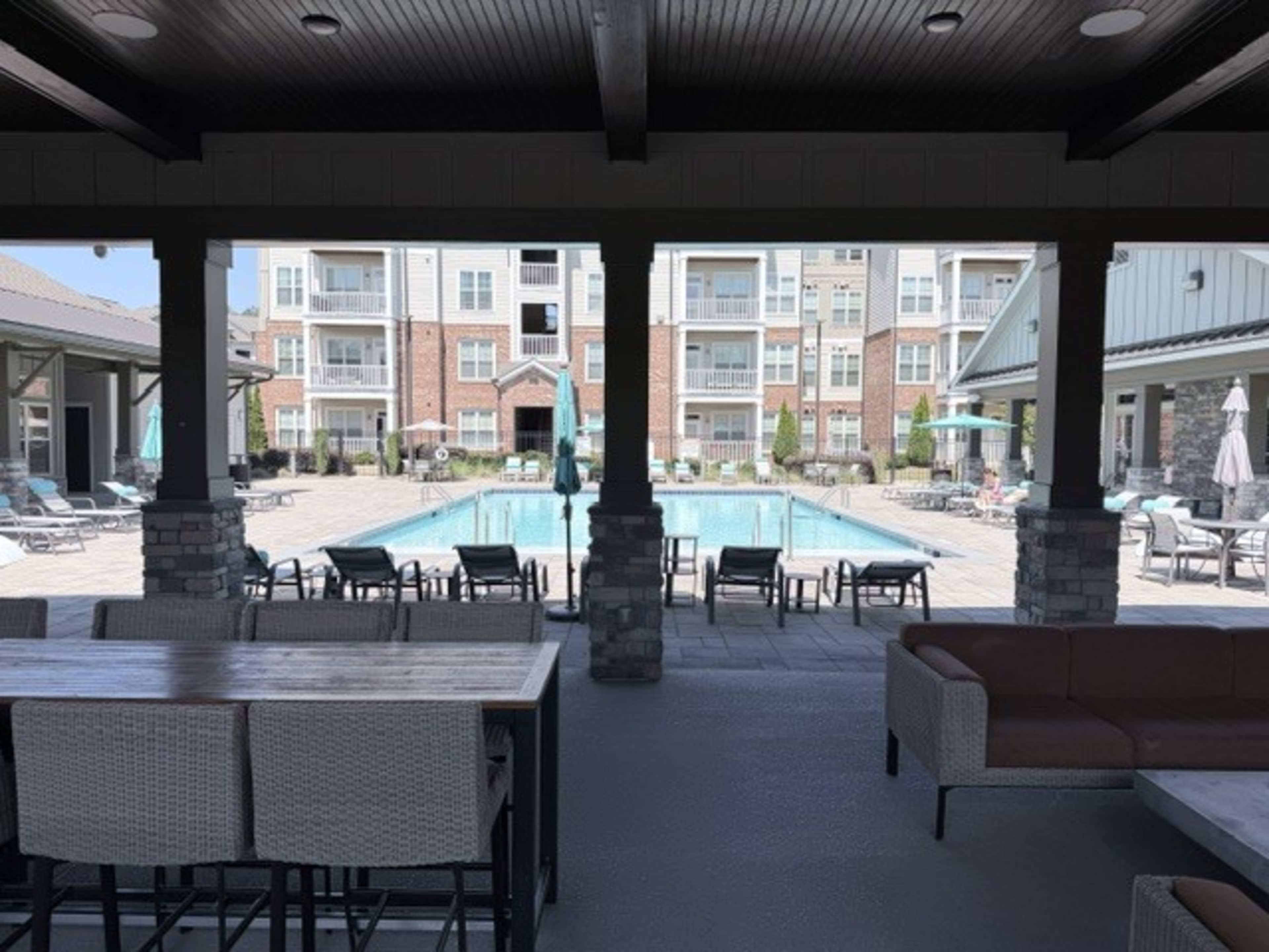 The image shows a covered outdoor area with seating and a view of a swimming pool surrounded by lounge chairs and apartment buildings in the background.