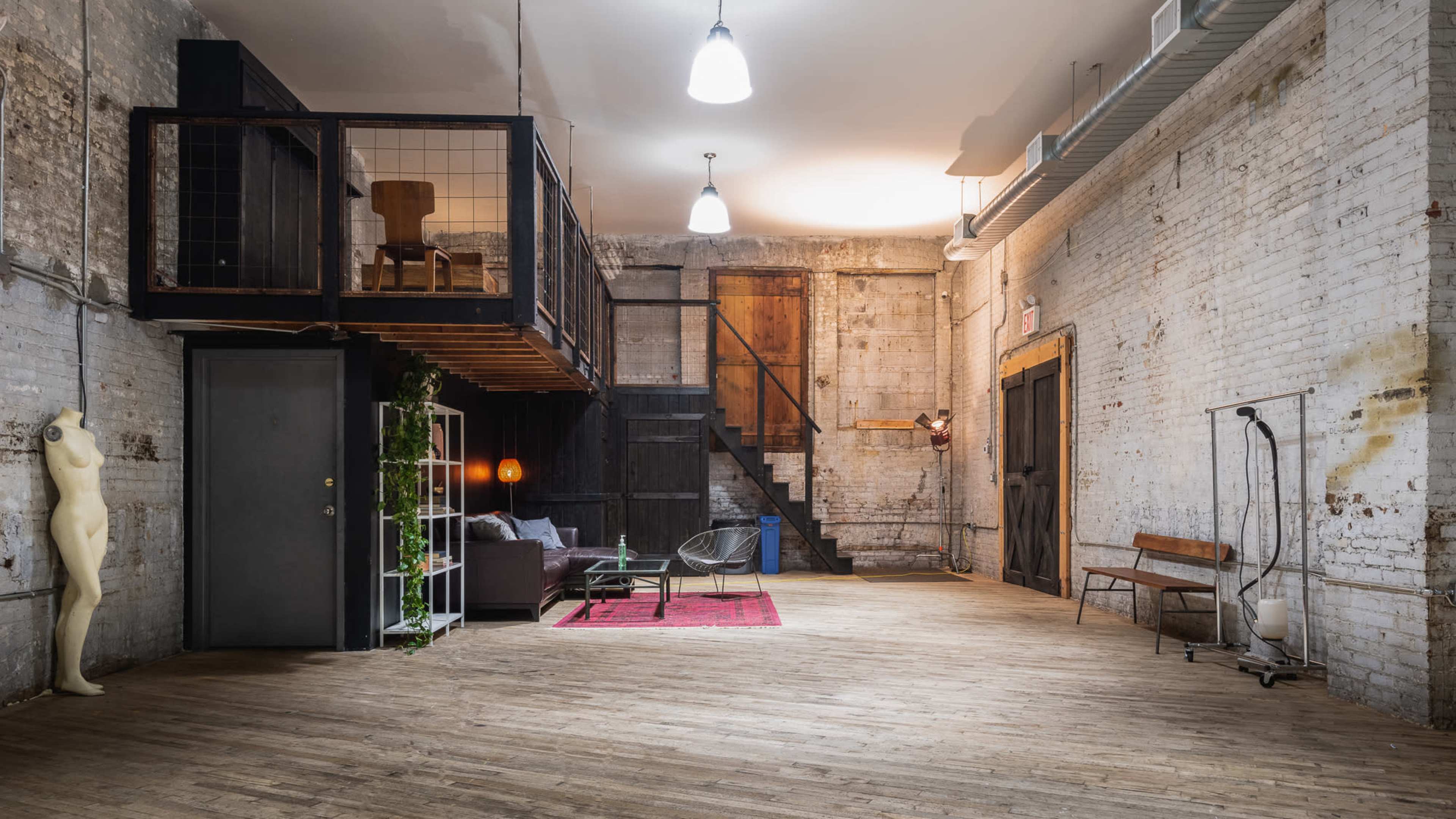The image shows a spacious, industrial-style room with a wooden floor, featuring a lofted area, a seating arrangement, and a mannequin in the corner.