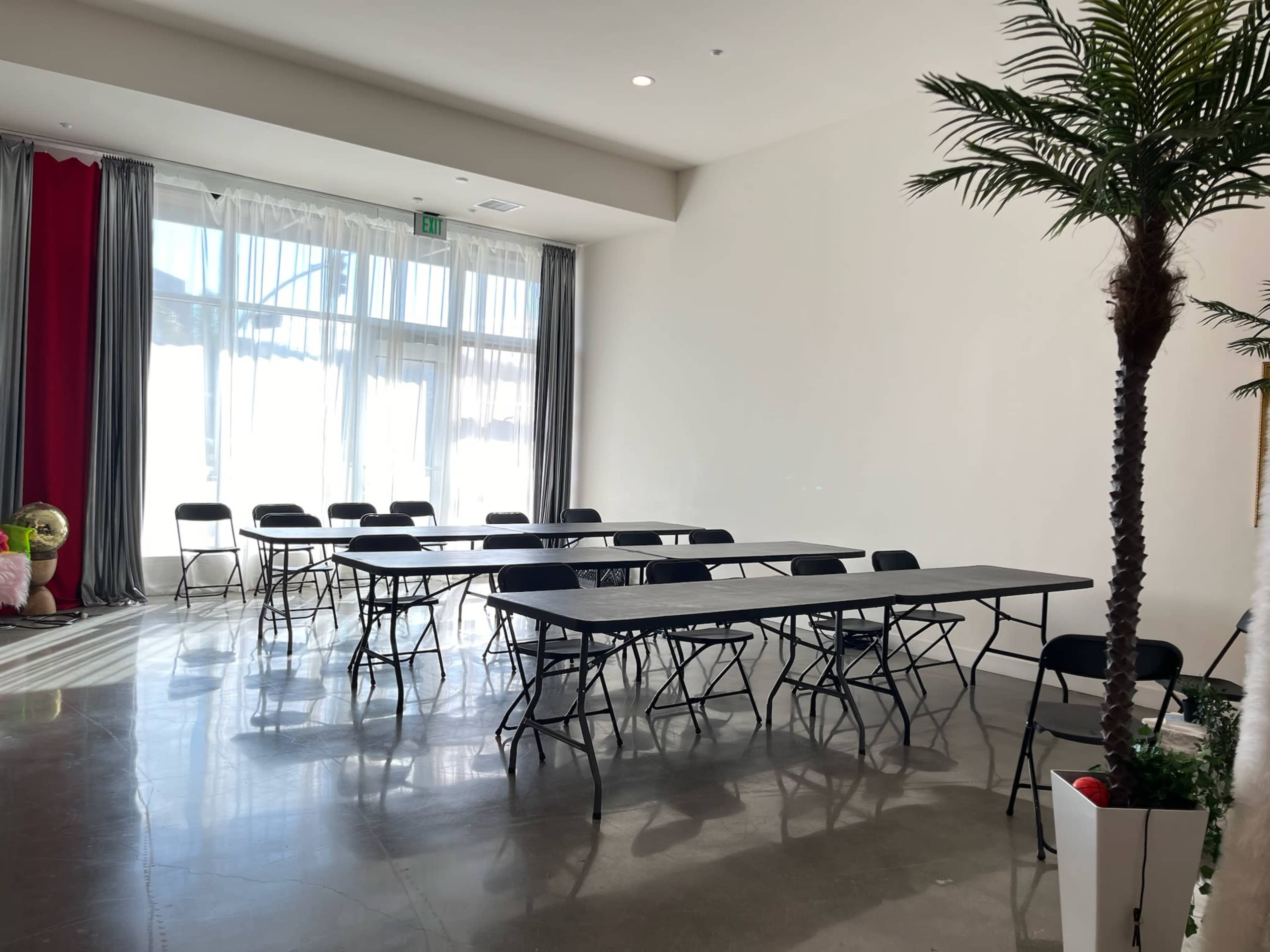 A bright room with multiple folding tables and chairs arranged for a meeting, featuring large windows and potted plants.