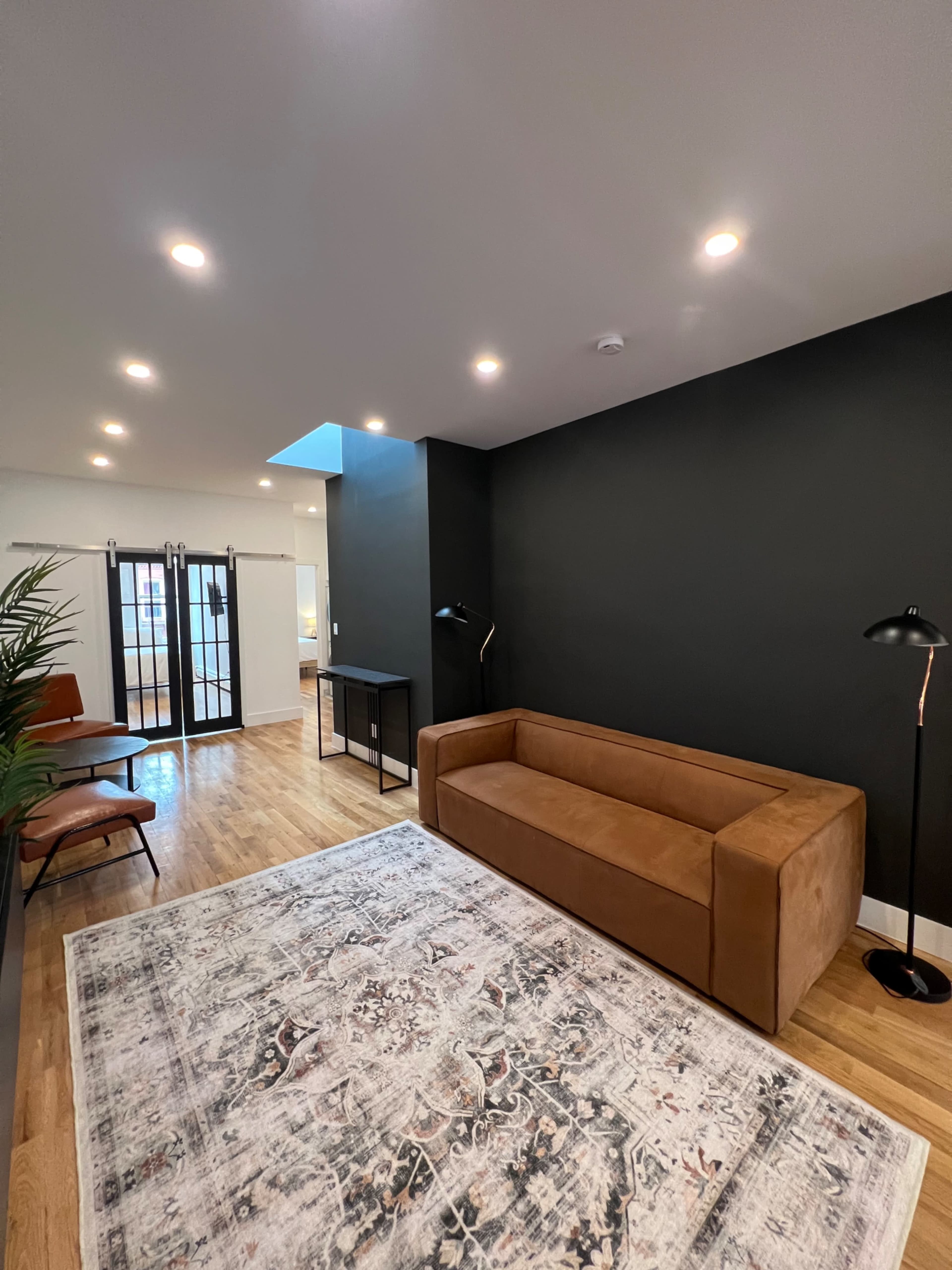 A modern living space features a brown sofa, a patterned rug, a standing lamp, and a sleek table against a dark wall.
