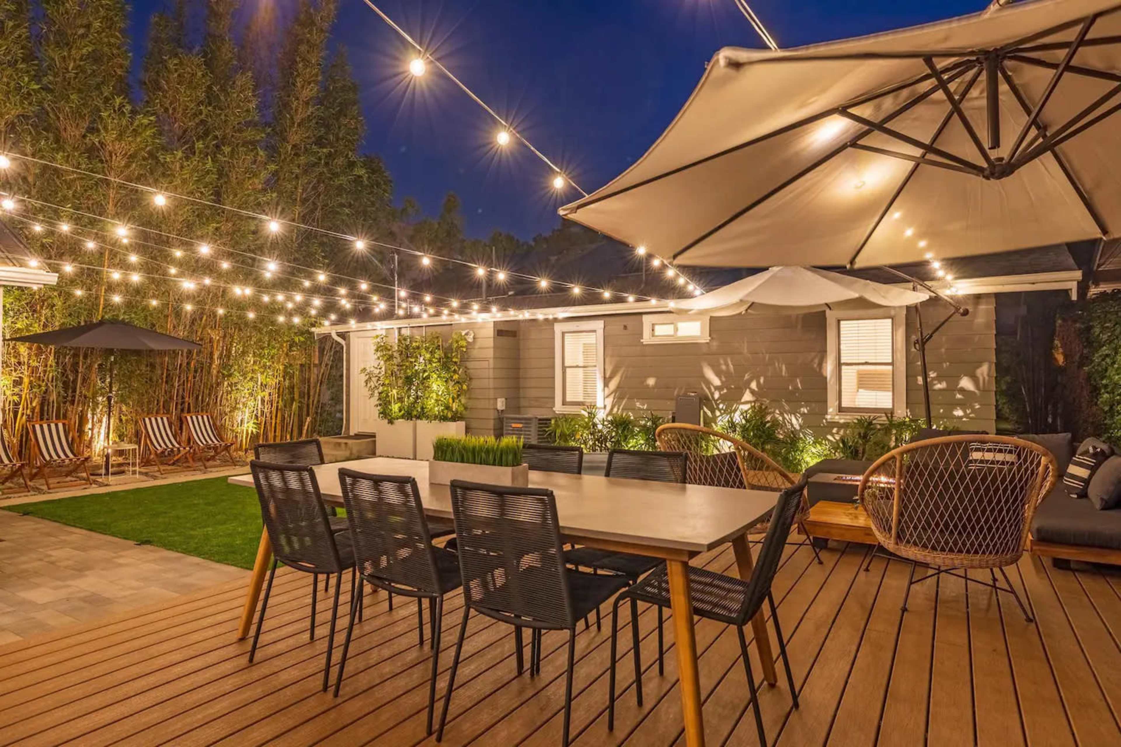 The image shows a backyard patio area featuring a wooden deck, a large dining table with chairs, and string lights overhead, set against a backdrop of landscaped greenery and a house.