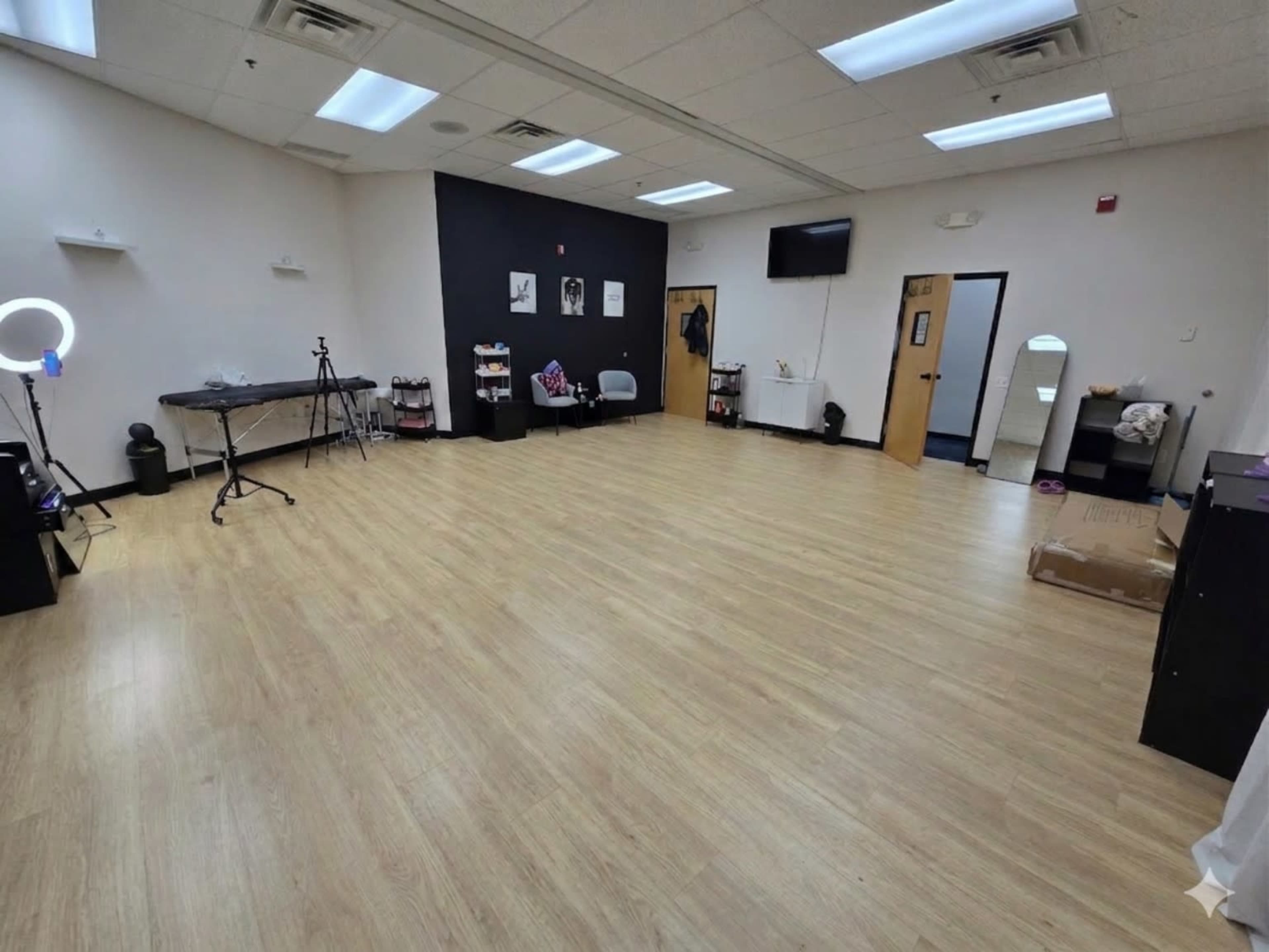 The image shows an empty studio space with light wood flooring, white and black walls, some furniture, and various equipment along the sides.