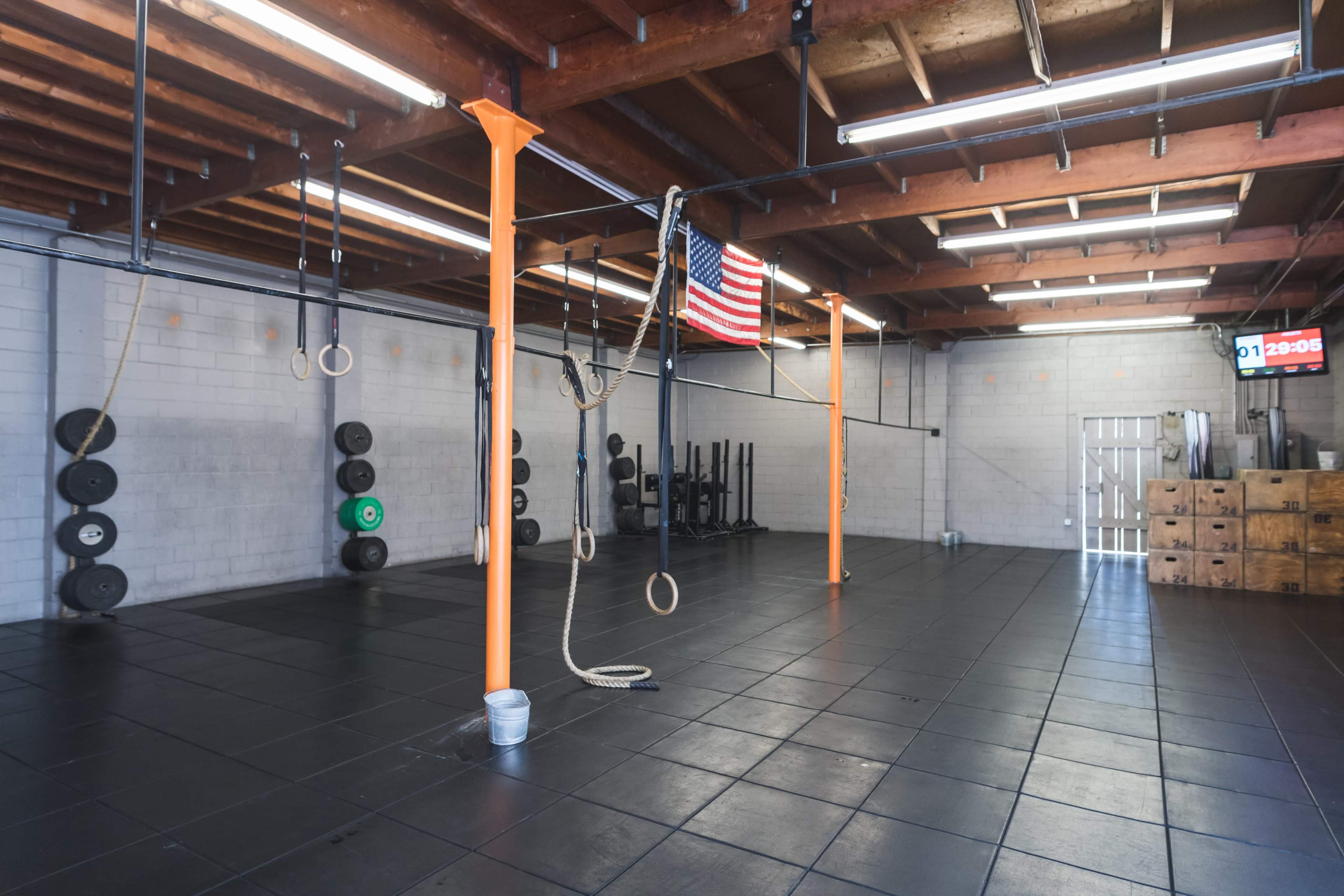 A spacious gym interior with black rubber flooring, weight plates stacked against the walls, and gymnastic rings hanging from overhead bars, an American flag displayed, and a clock showing the time.