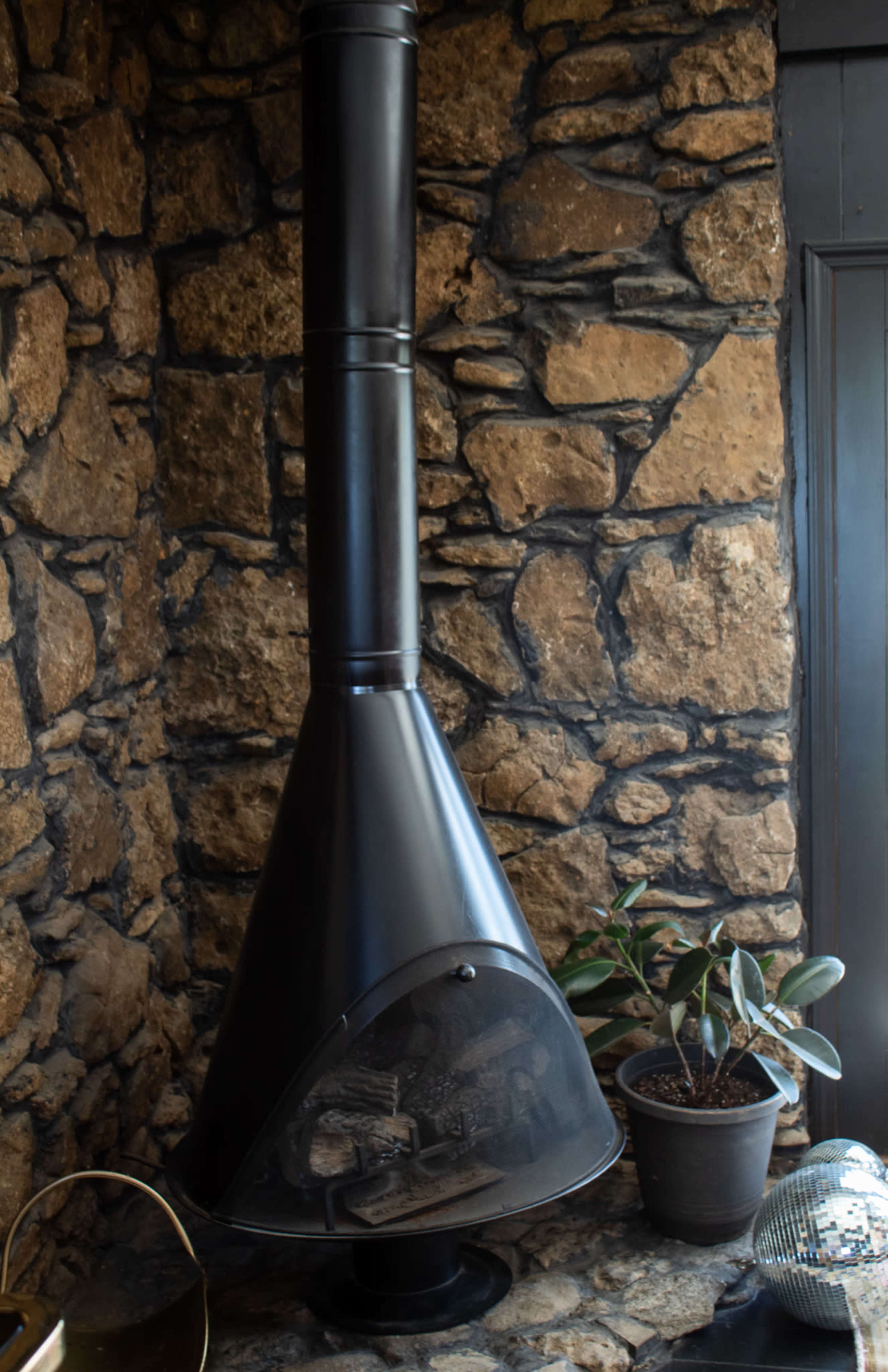 A black, modern wood-burning stove is positioned against a textured stone wall, with a potted plant nearby.