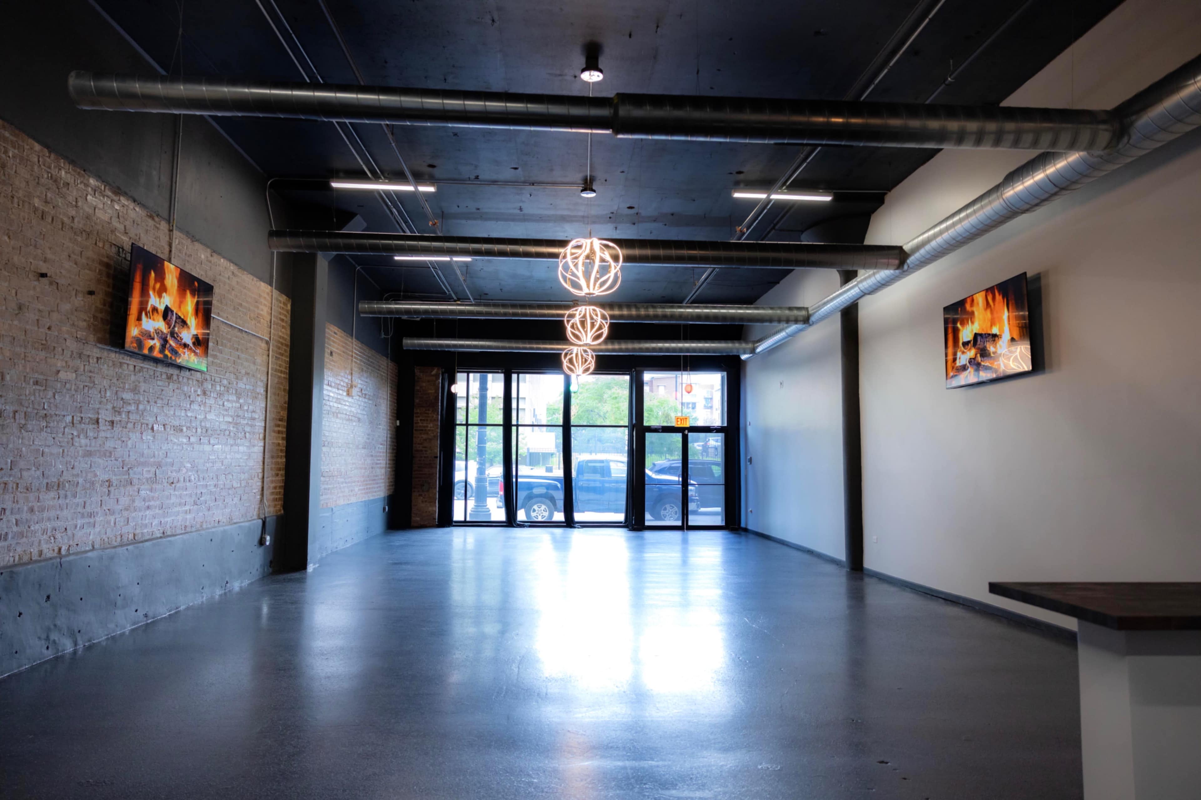 The image shows an empty commercial space with a polished concrete floor, large windows at the entrance, and two mounted screens displaying images of fireplaces.