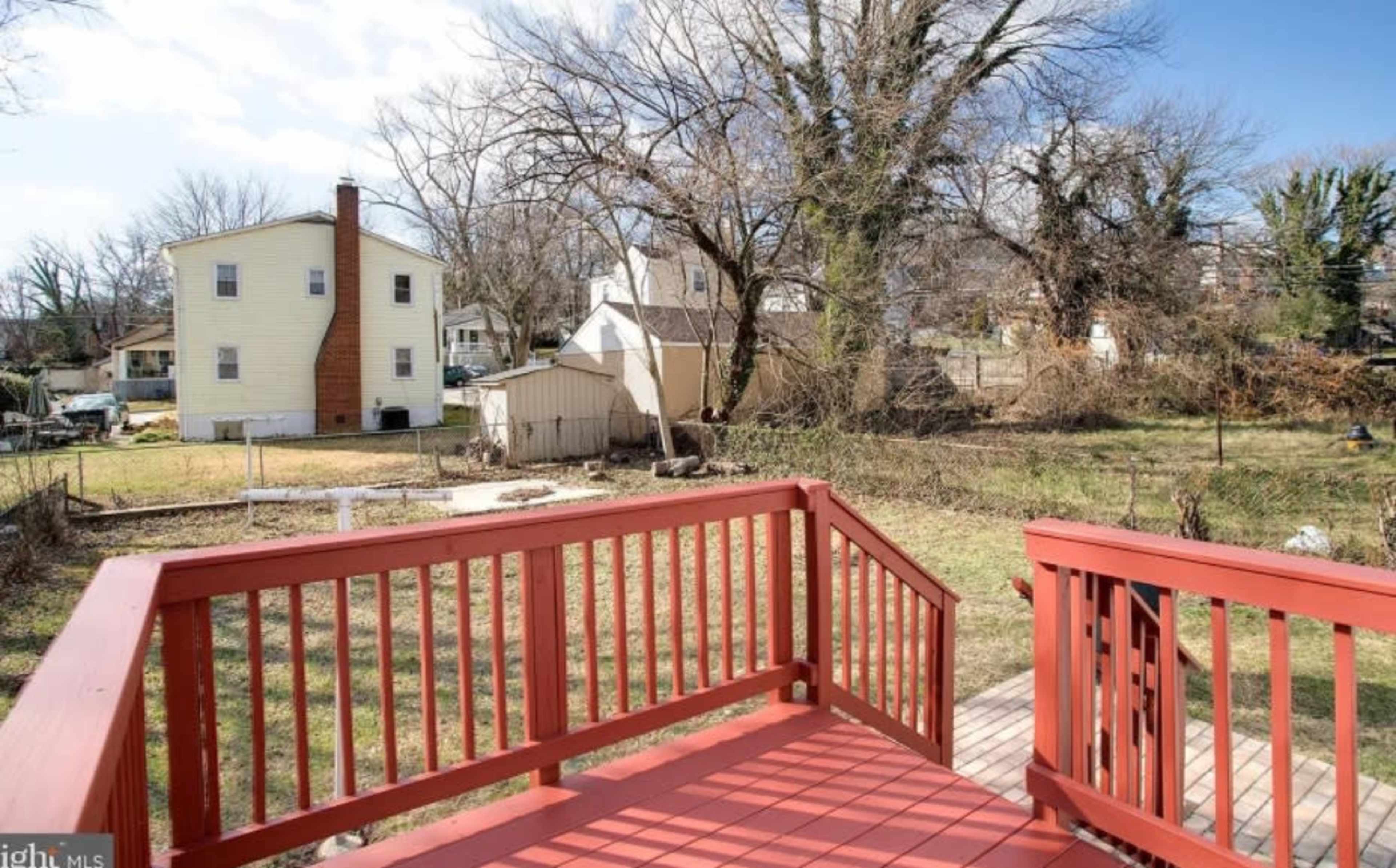 A wooden deck overlooks a grassy yard with several trees and two houses in the background.