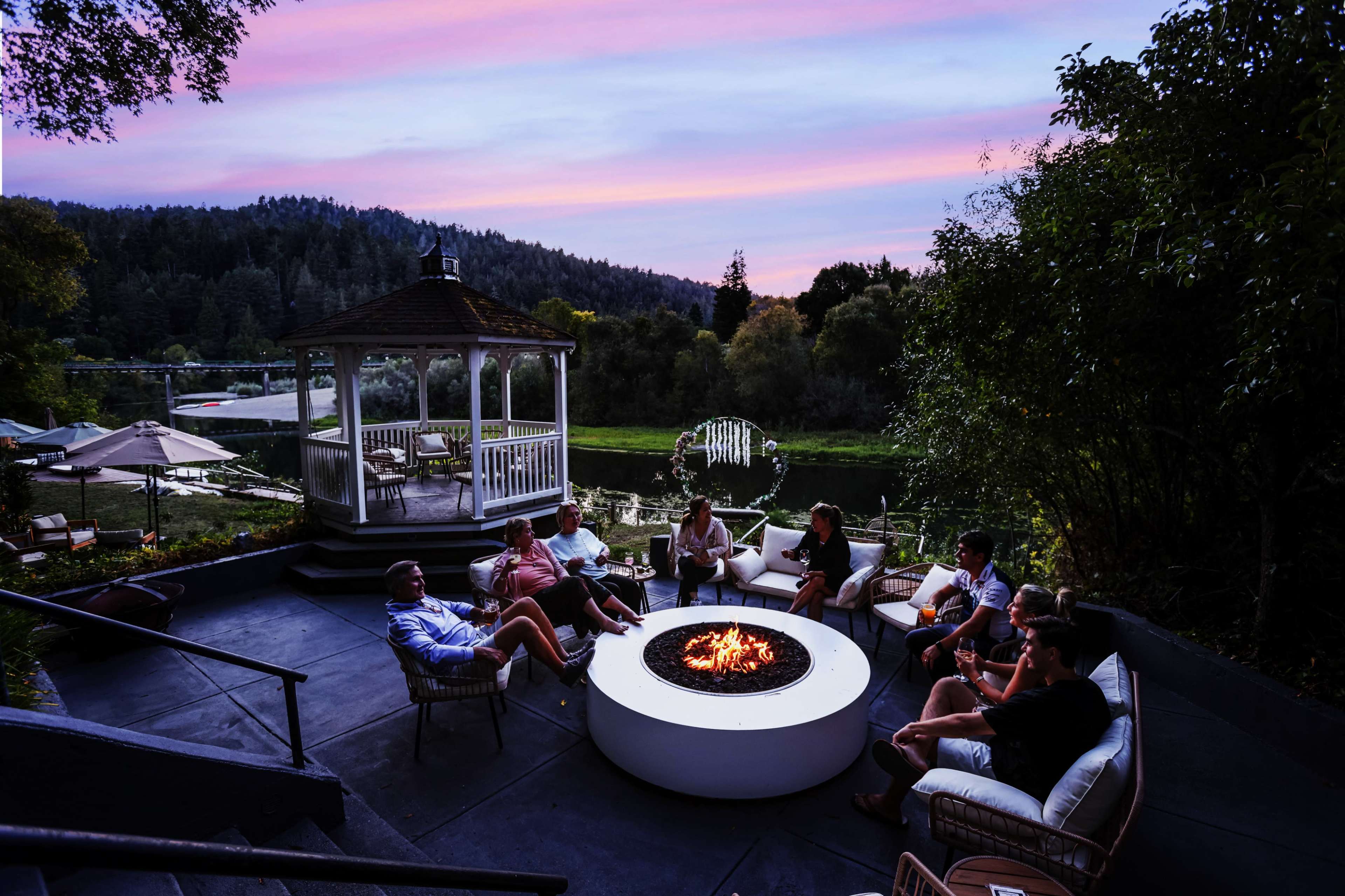 A group of people sits around a circular fire pit in an outdoor setting, with a gazebo and wooded area in the background at sunset.