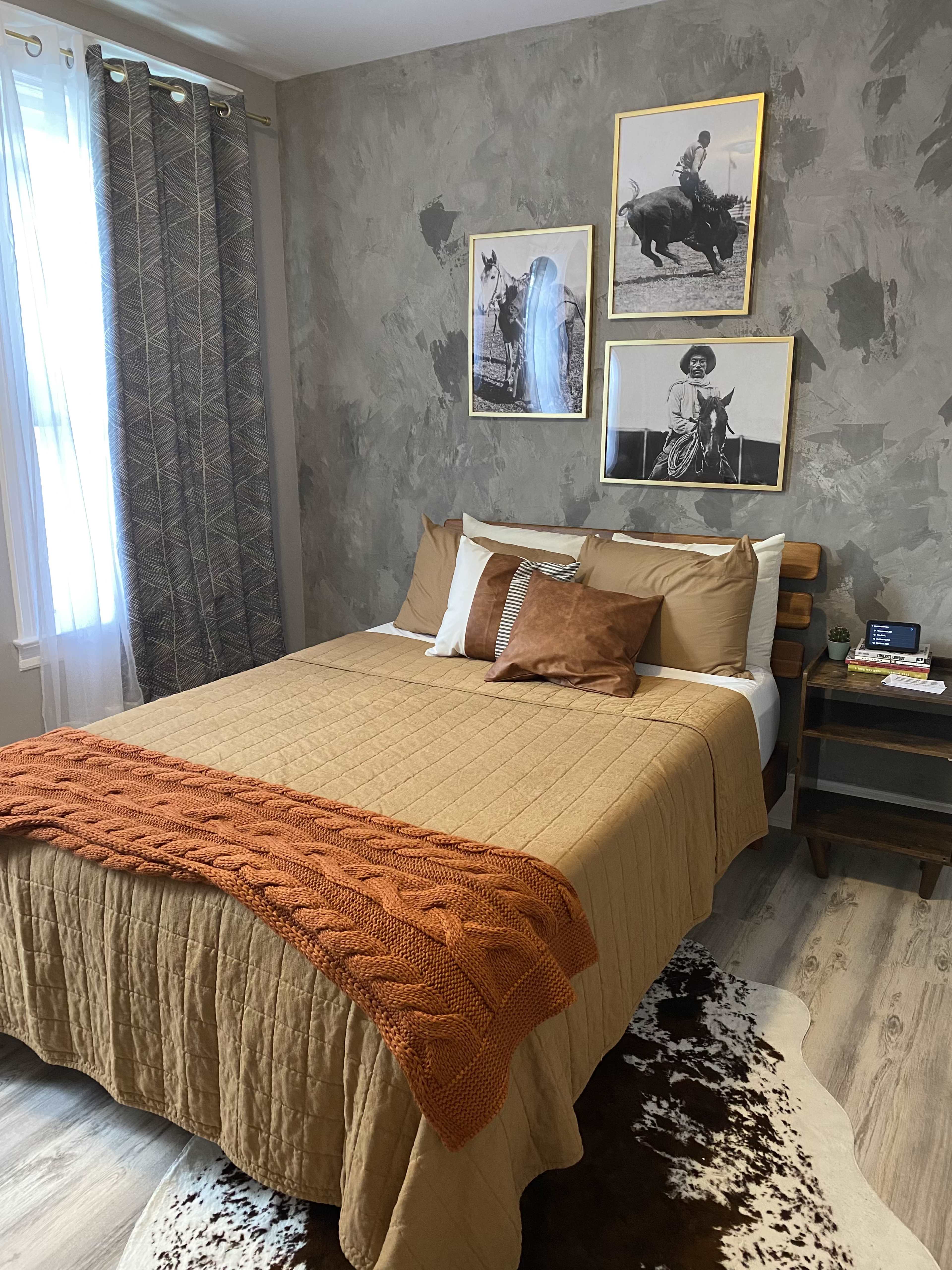 The image shows a bedroom with a queen-sized bed covered in a beige quilt, accented by orange pillows and a knitted throw, flanked by framed photographs on the gray textured walls.