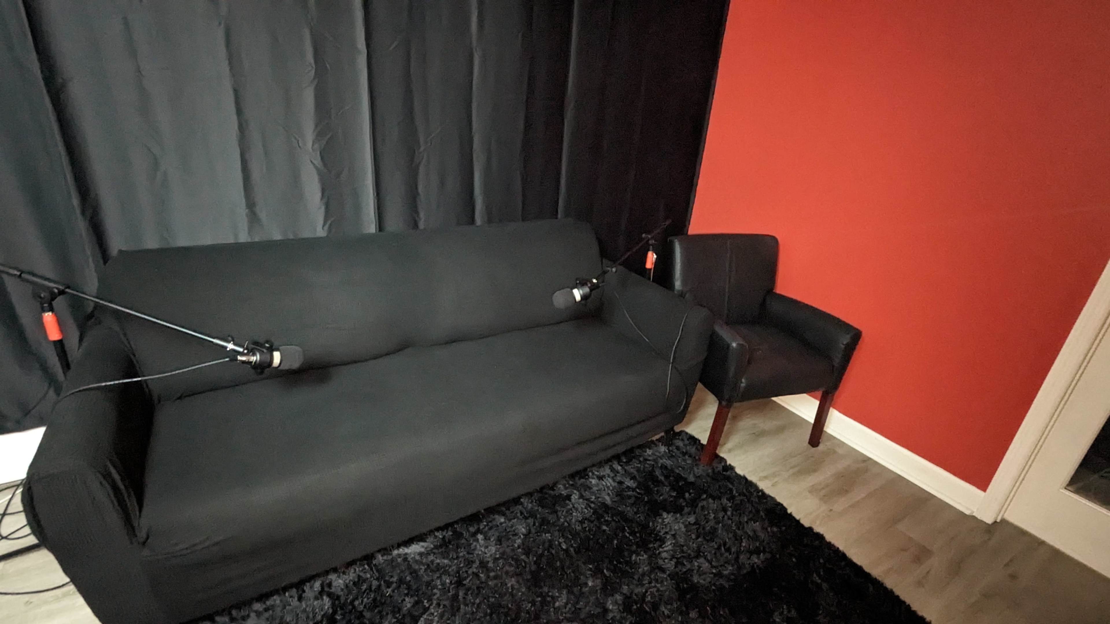 A black couch and a black armchair are positioned against a red wall, with microphones mounted on both chairs.