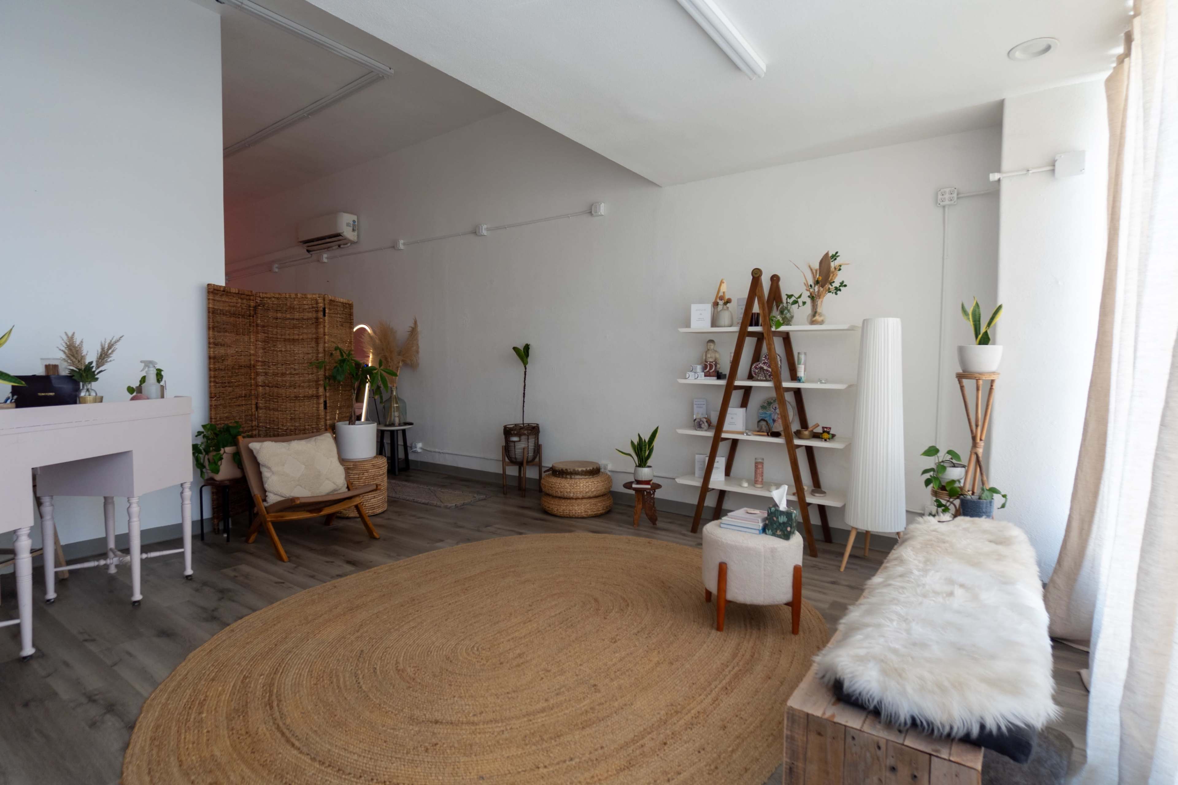 The image shows a minimalist room with wooden floors, a round natural fiber rug, a seating area with a chair and ottoman, and shelves displaying decorative items and plants.