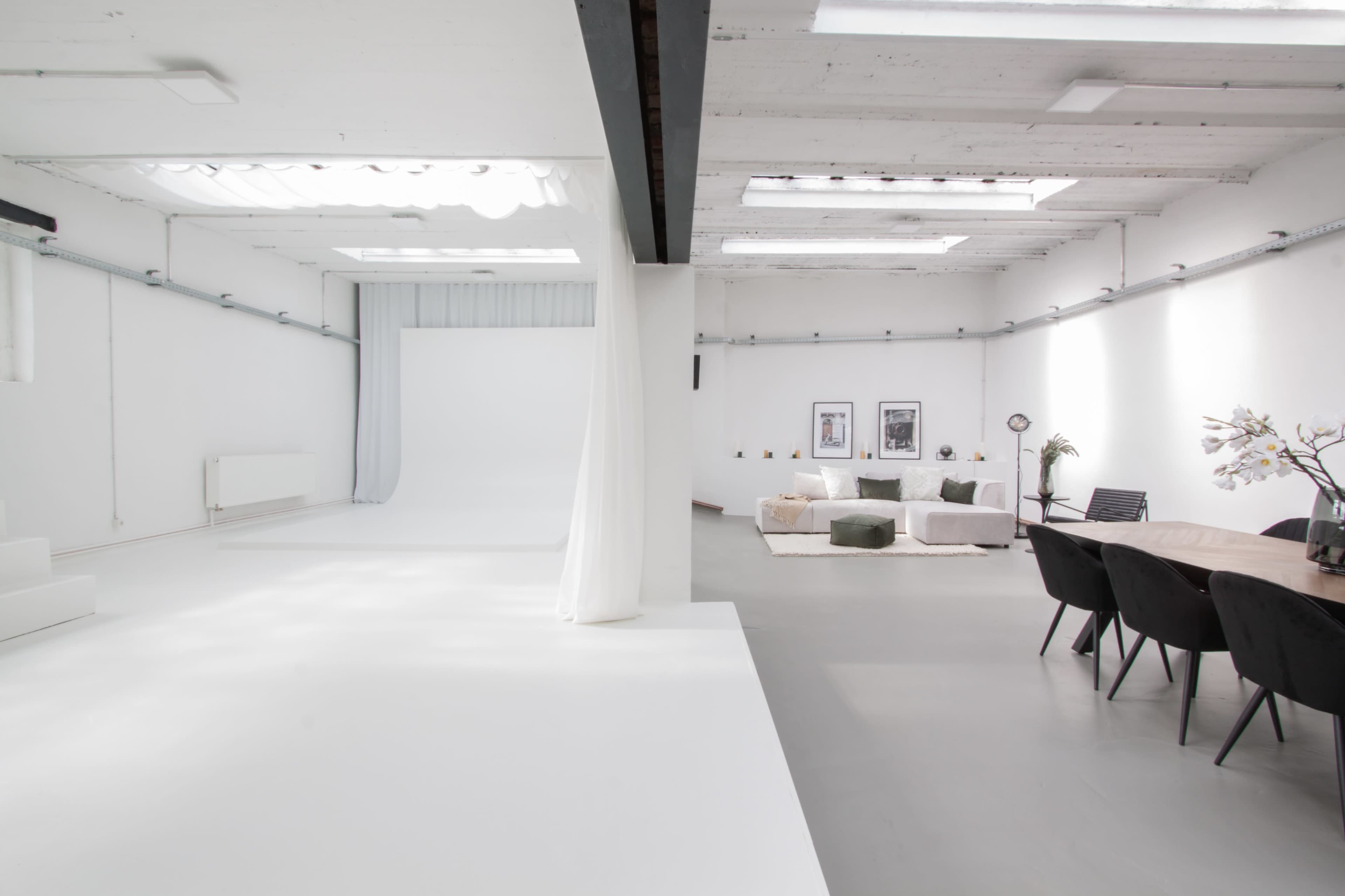 A bright, spacious studio featuring a white backdrop on one side and a cozy seating area with a dining table on the other.