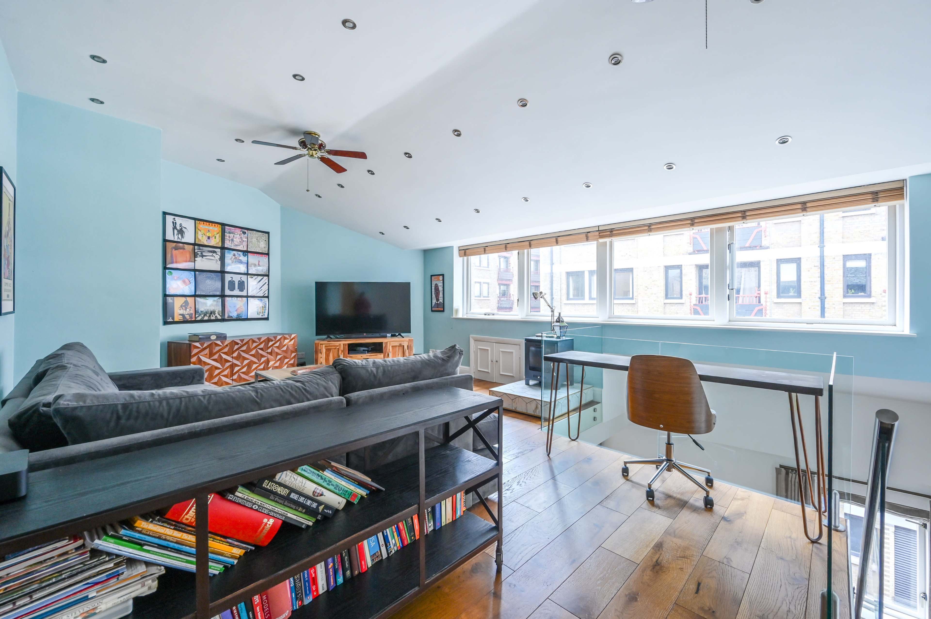 The room features a light blue wall with a sloped ceiling, a sofa, a television, a desk with a chair, and shelves filled with books.