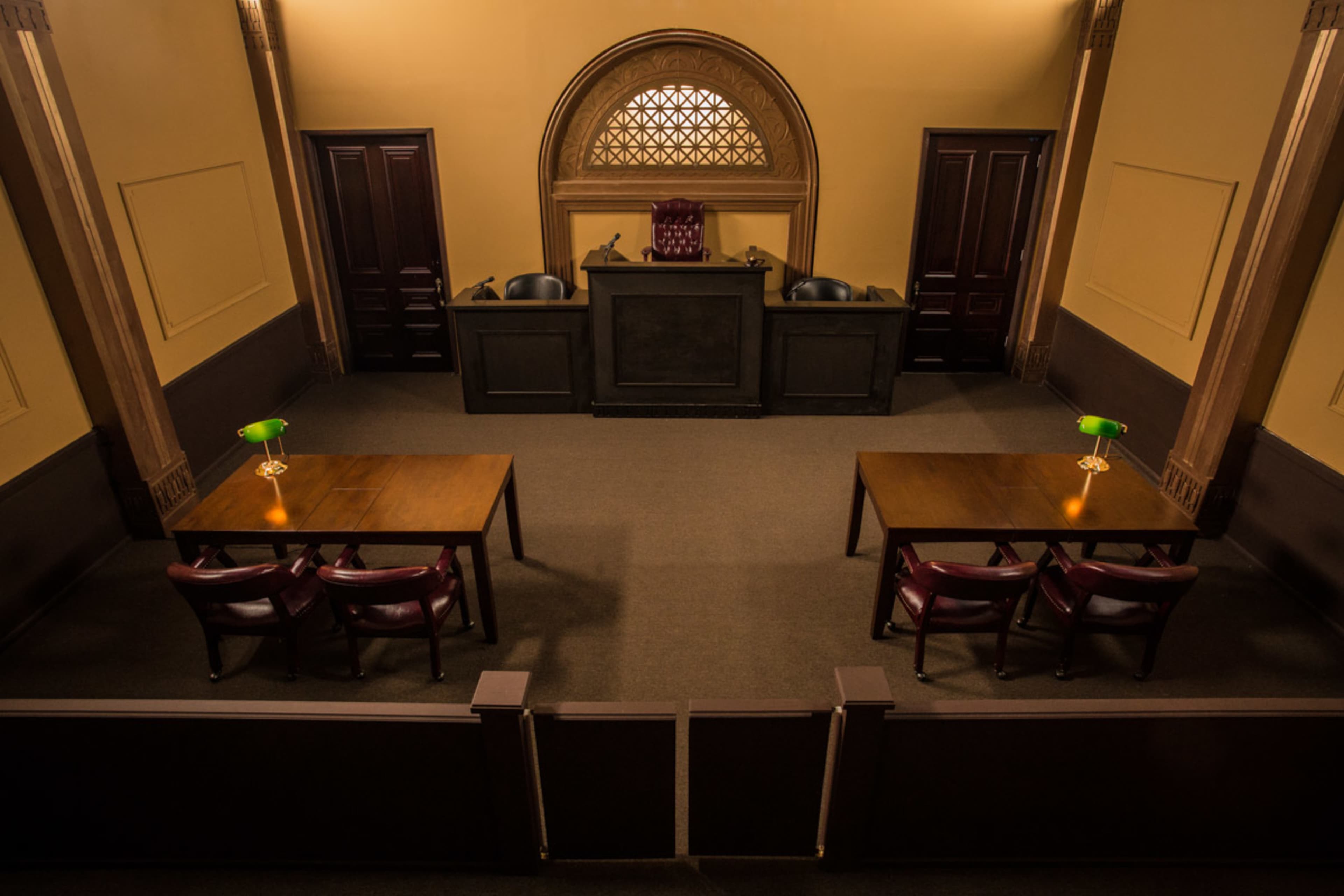 The image shows a courtroom interior with two wooden tables and chairs positioned in front of a judge's bench, lit by green desk lamps.
