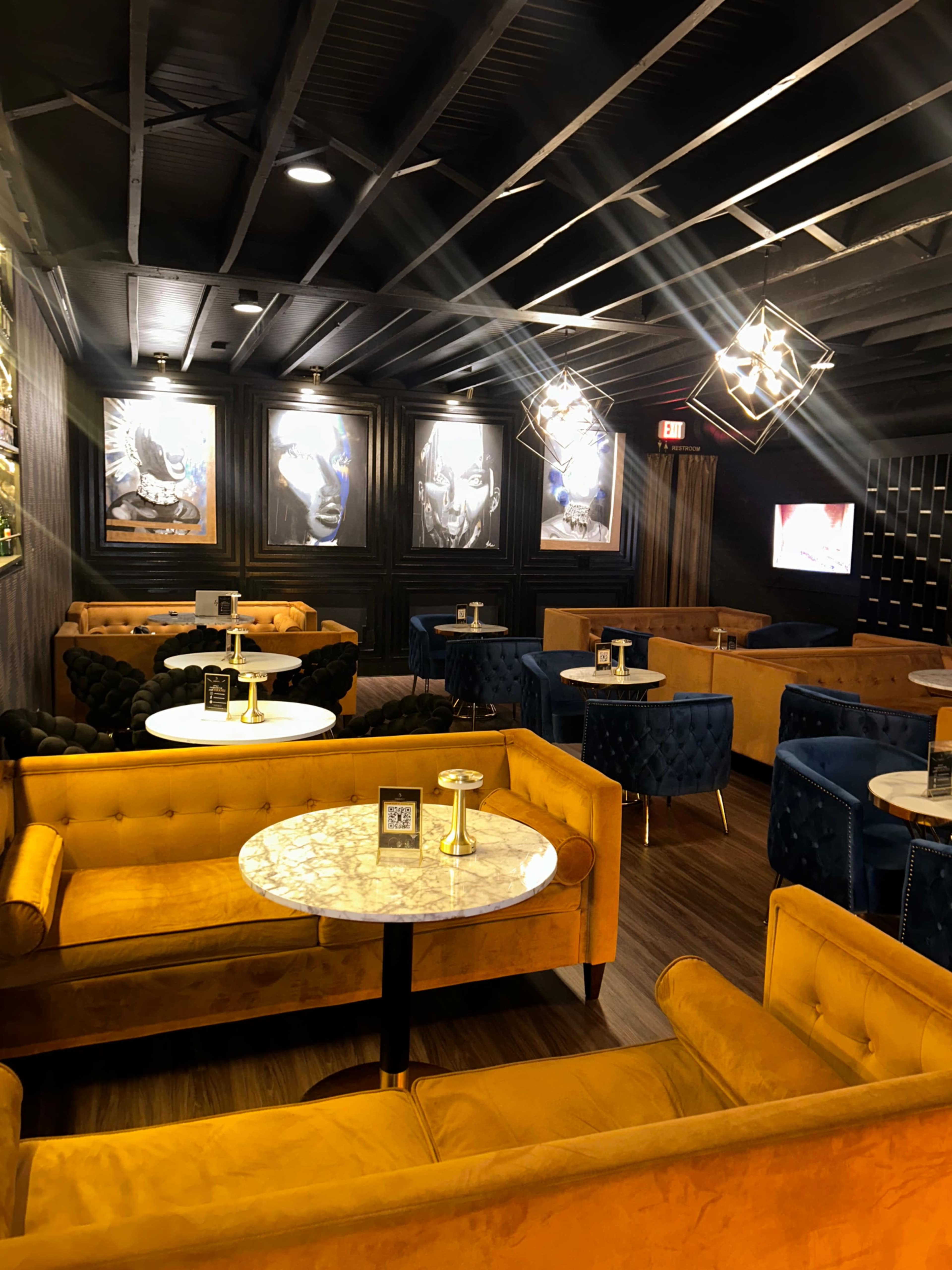 The image shows a modern lounge with yellow and blue upholstered seating, circular marble-topped tables, and framed artwork on the walls.