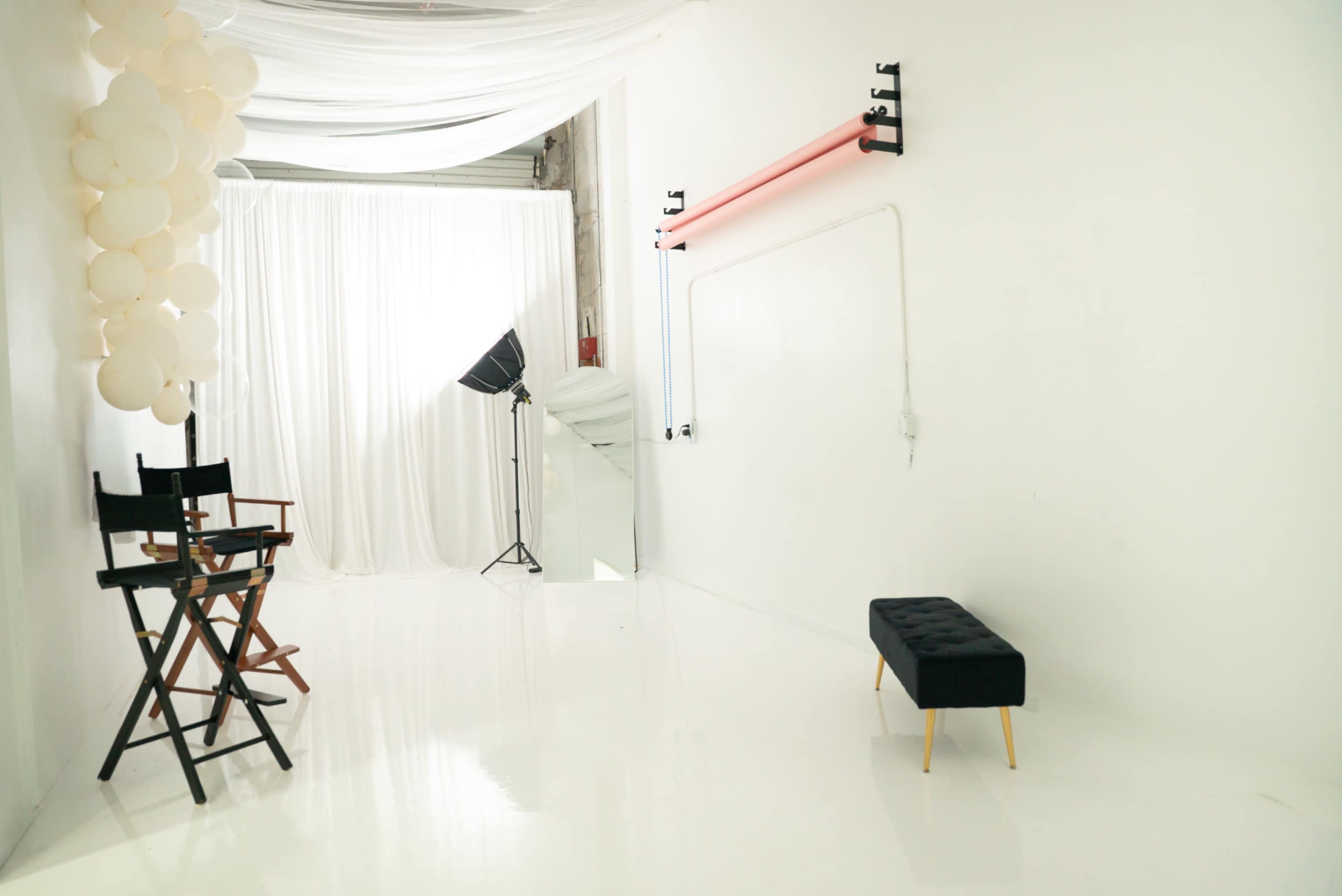 The image shows a spacious, minimalist studio with a white backdrop, a director's chair, a black bench, and a photography light setup.