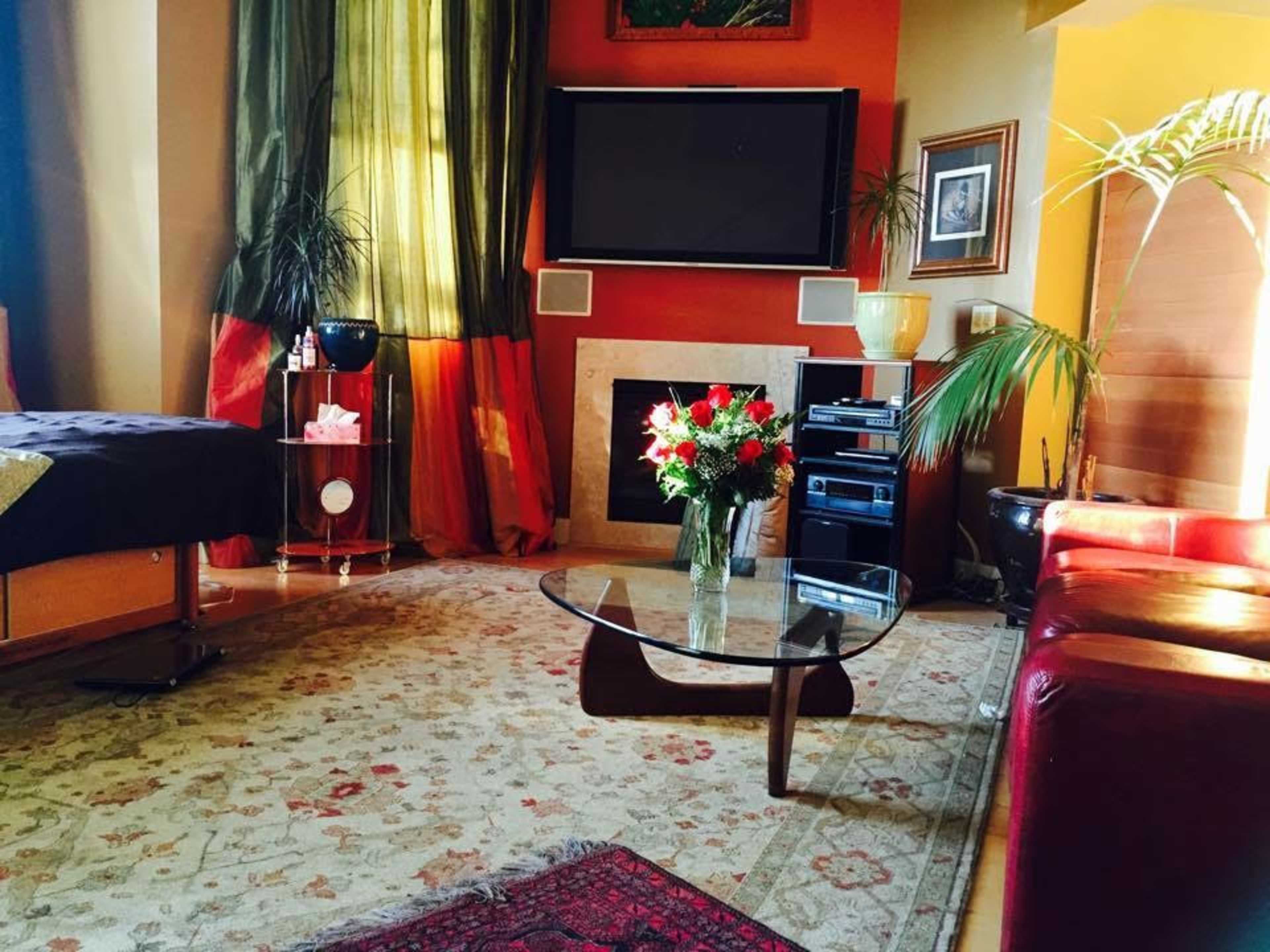 A living room with a glass coffee table, a floral arrangement, and a television mounted on the wall, surrounded by decorative plants and warm-colored curtains.