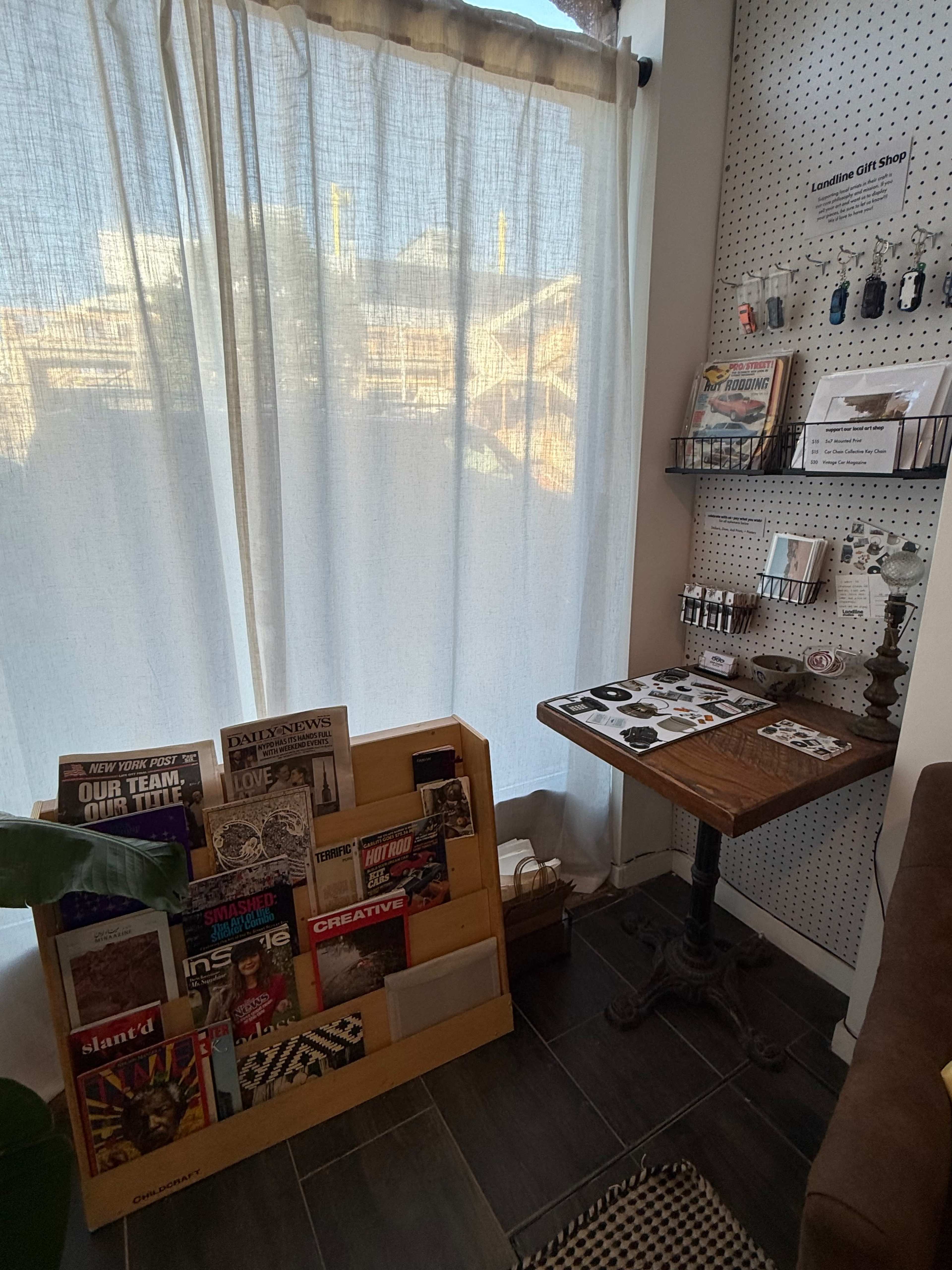 The image shows a small indoor space with a wooden magazine rack displaying various publications, a table with items for sale, and a white curtain partially covering a window.
