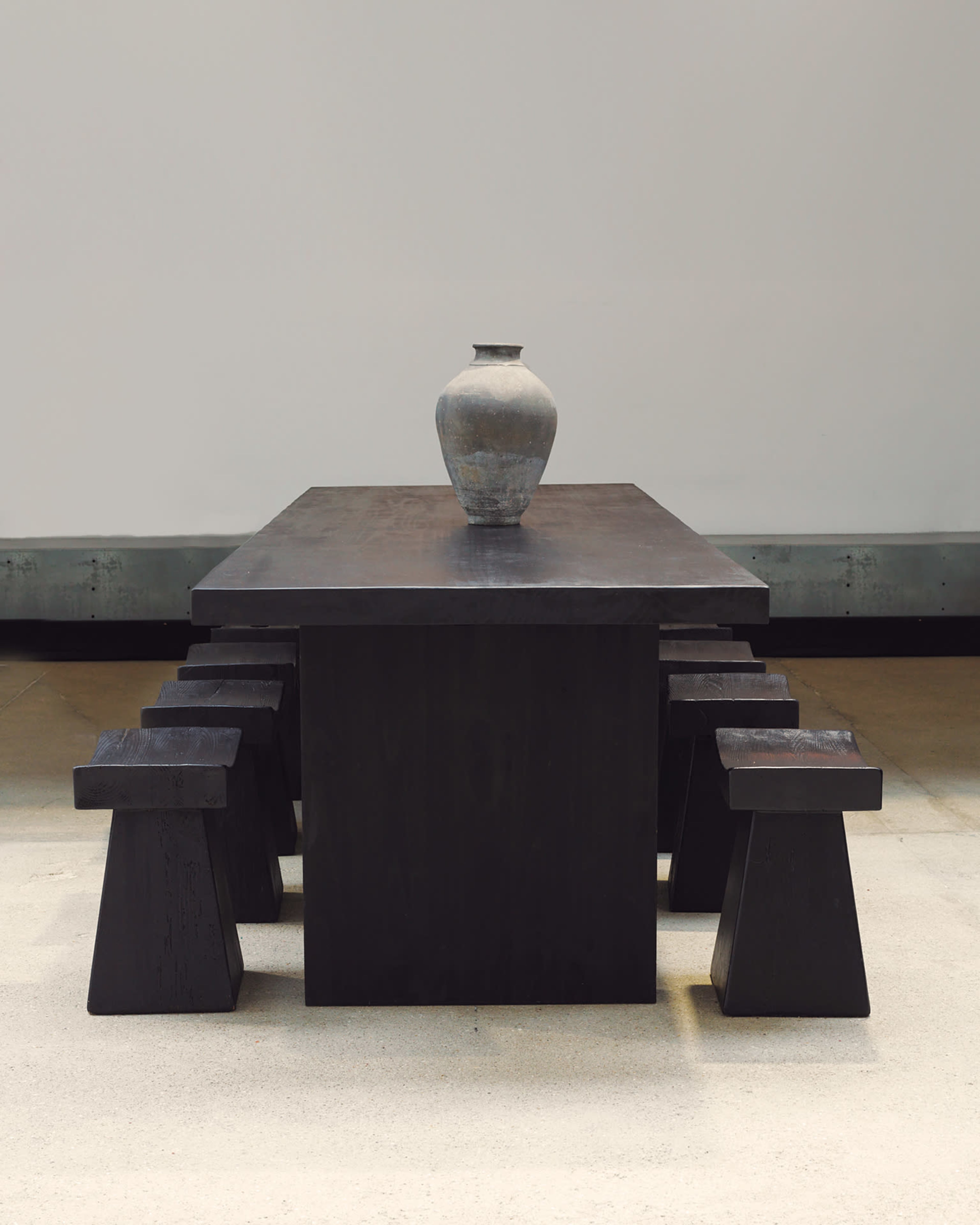 A rectangular black table is positioned in a spacious area with a ceramic vase placed in the center, surrounded by four matching black stools.
