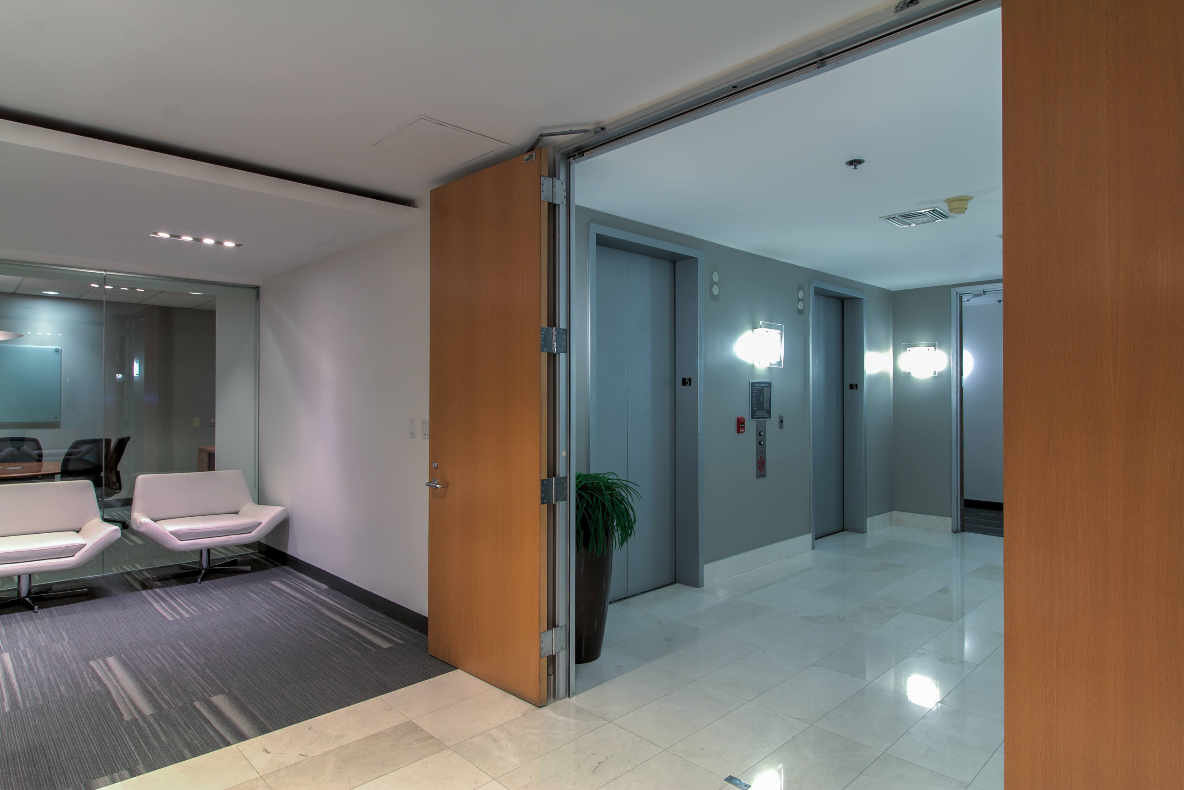 A modern office lobby with an open doorway leading to an elevator area and a seating area to the left.