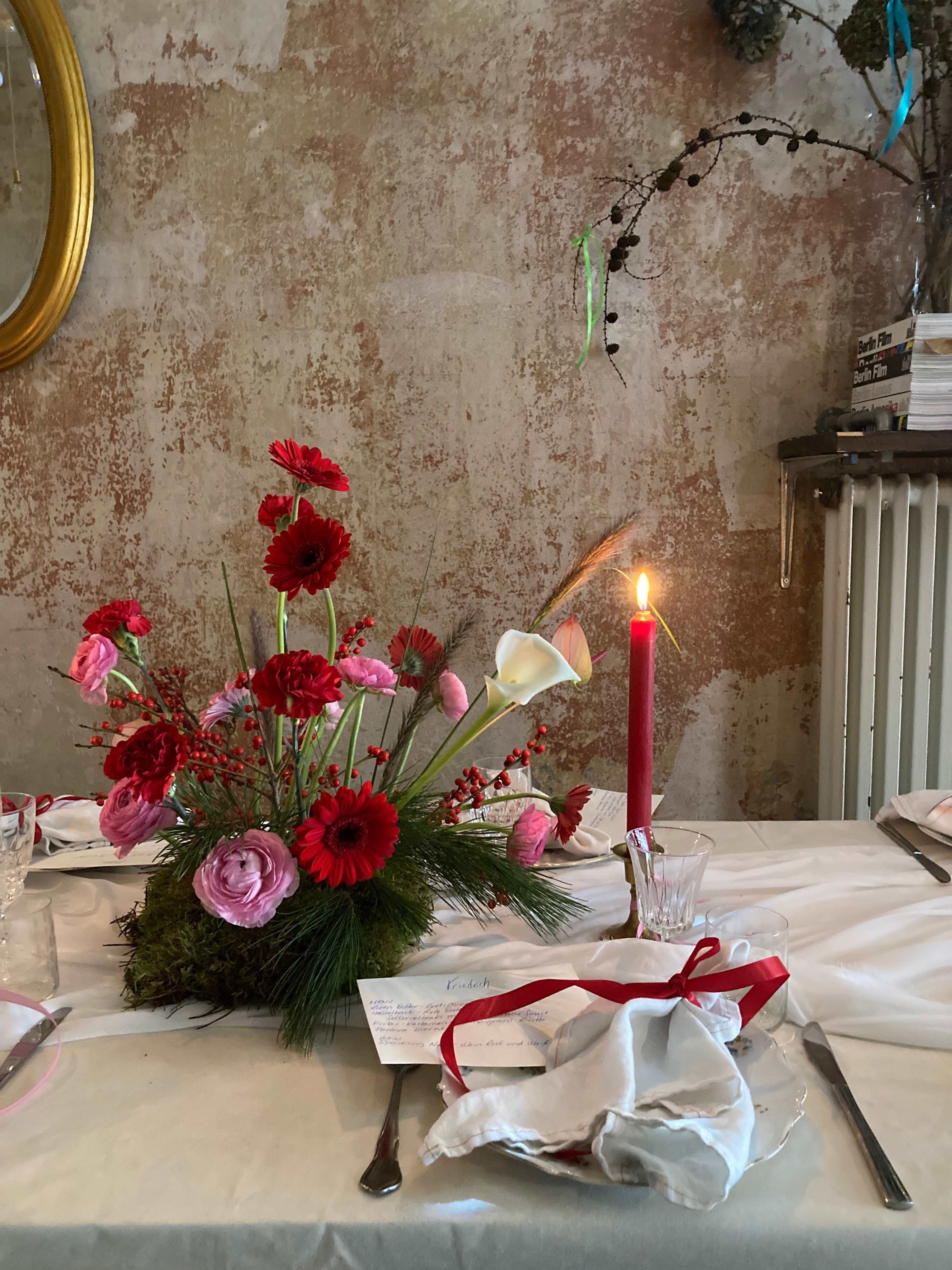 A table is set with a floral centerpiece featuring red and pink flowers, a lit candle, and neatly arranged silverware and napkins.
