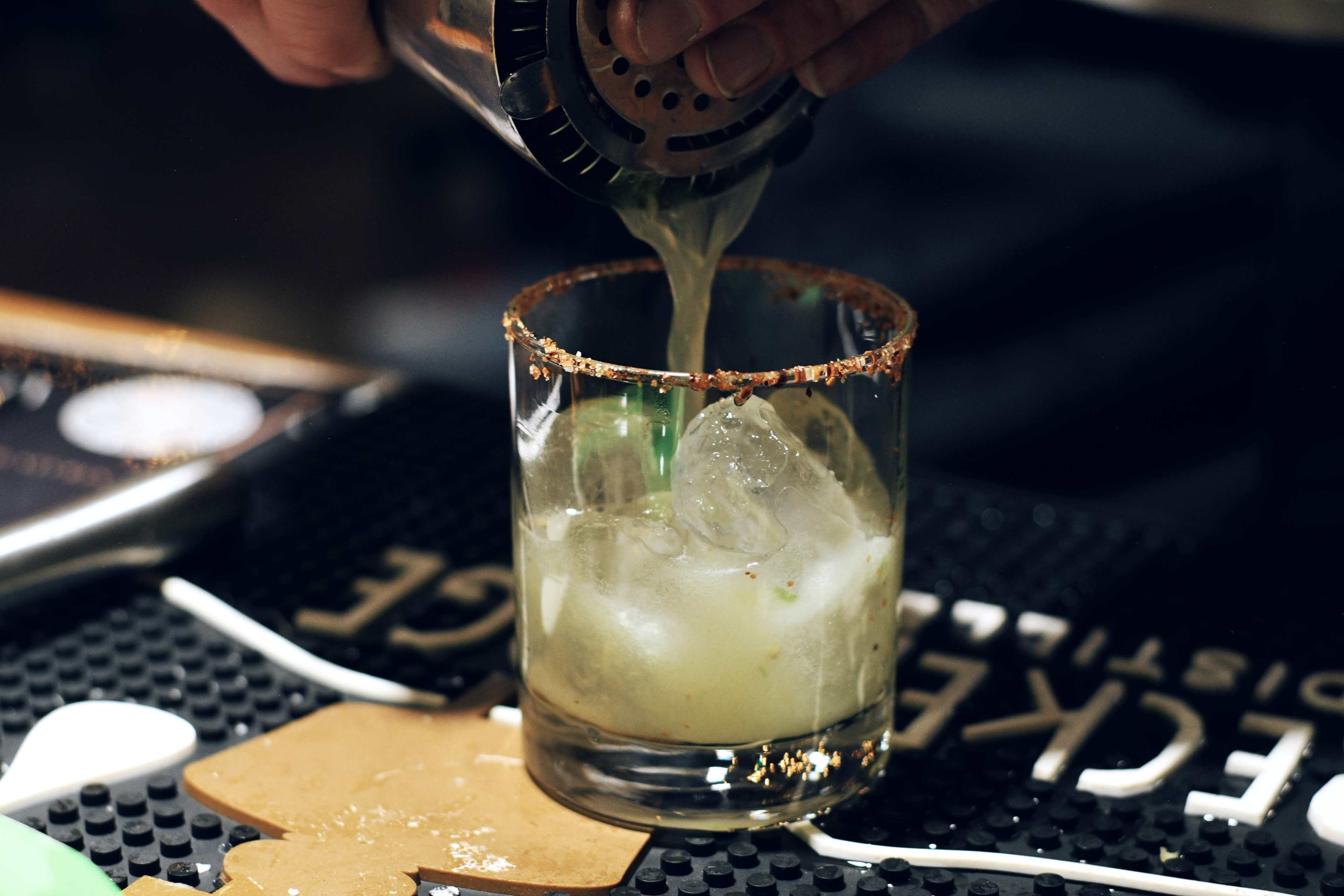 A bartender pours a cocktail from a shaker into a glass filled with ice and garnished with a salted rim.