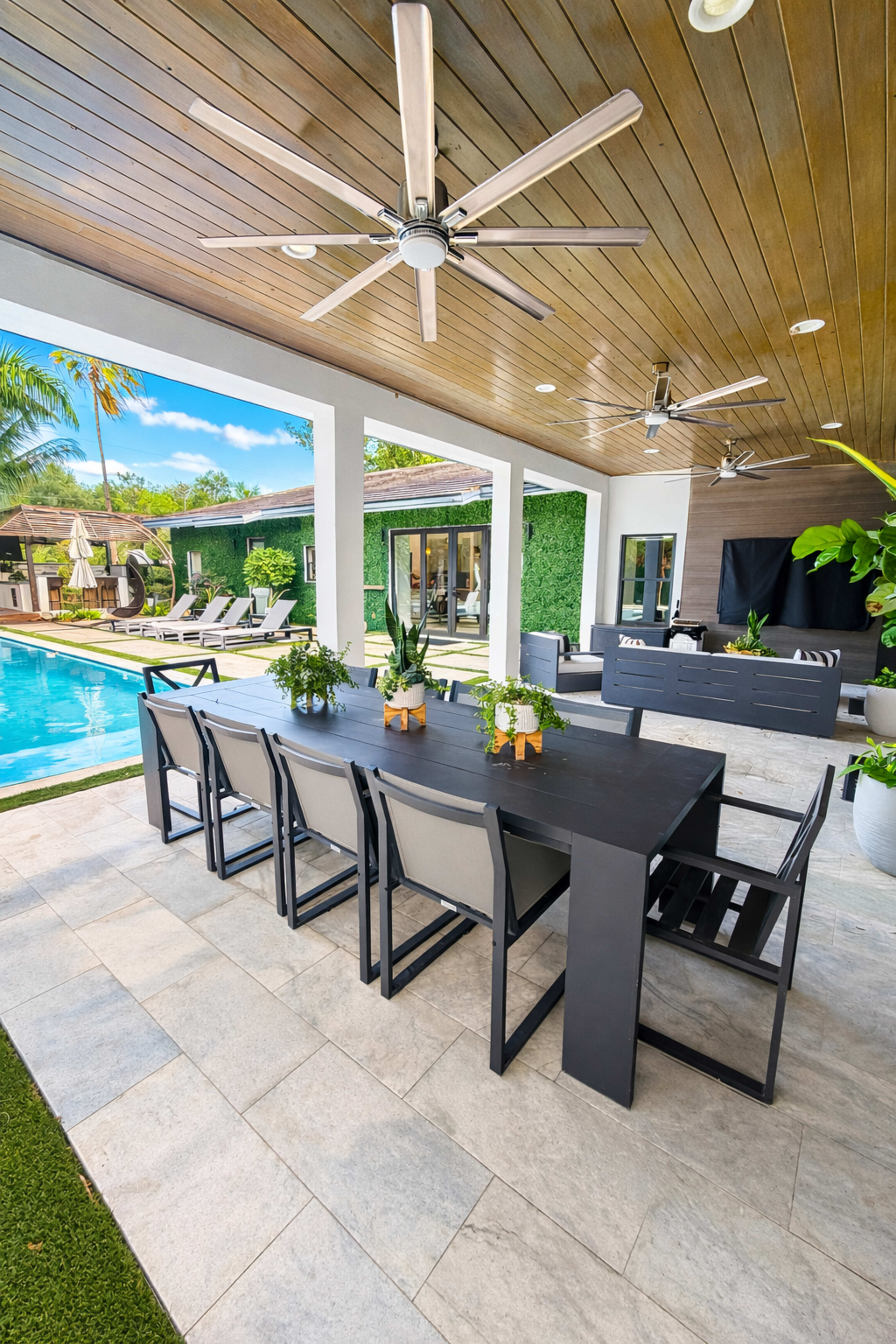 The image shows an outdoor dining area with a large table set next to a swimming pool, surrounded by tropical plants and sleek lounge chairs.