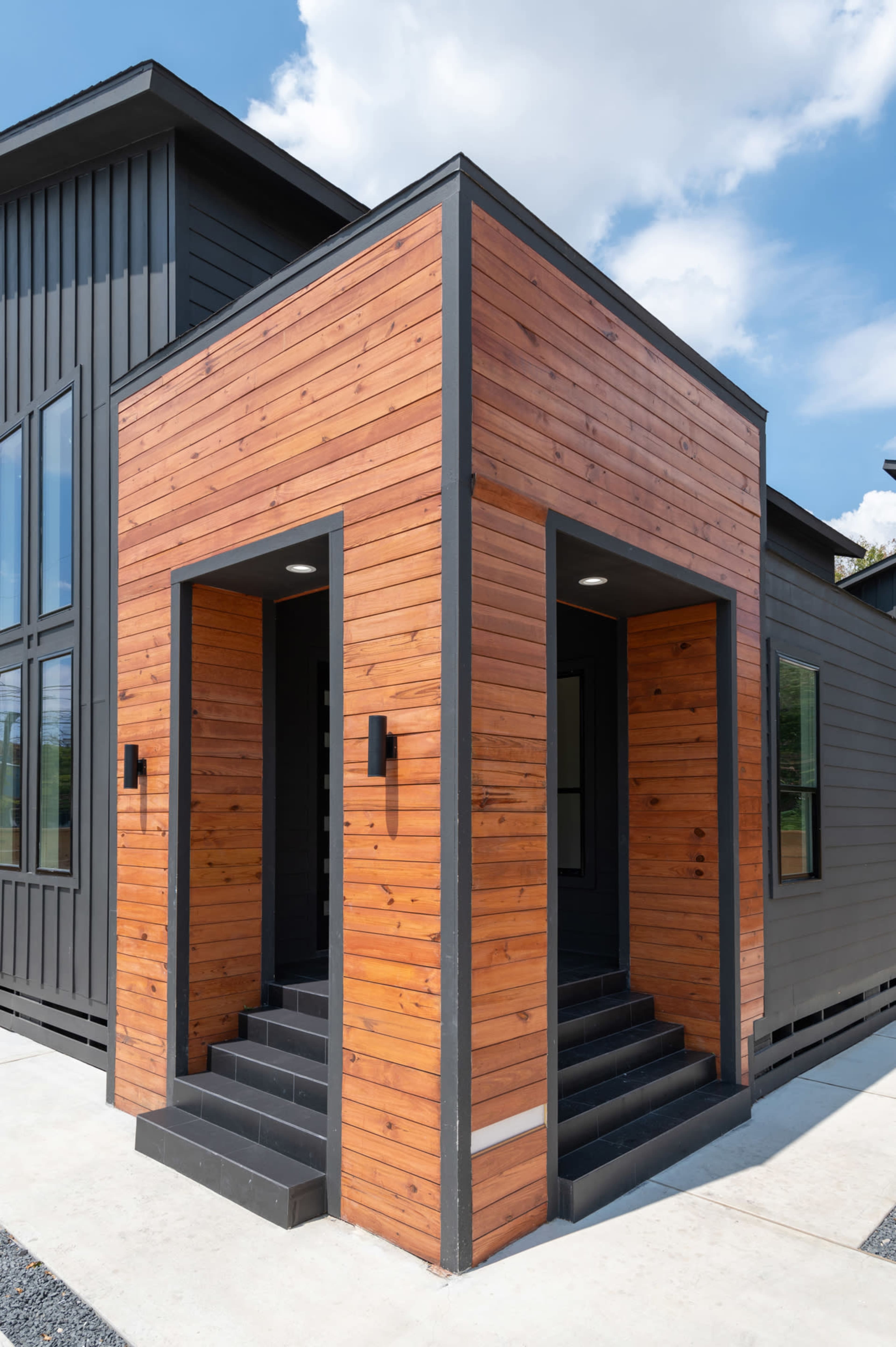 The image shows a modern house with a wooden exterior and two entrance stairways, set against a blue sky with scattered clouds.