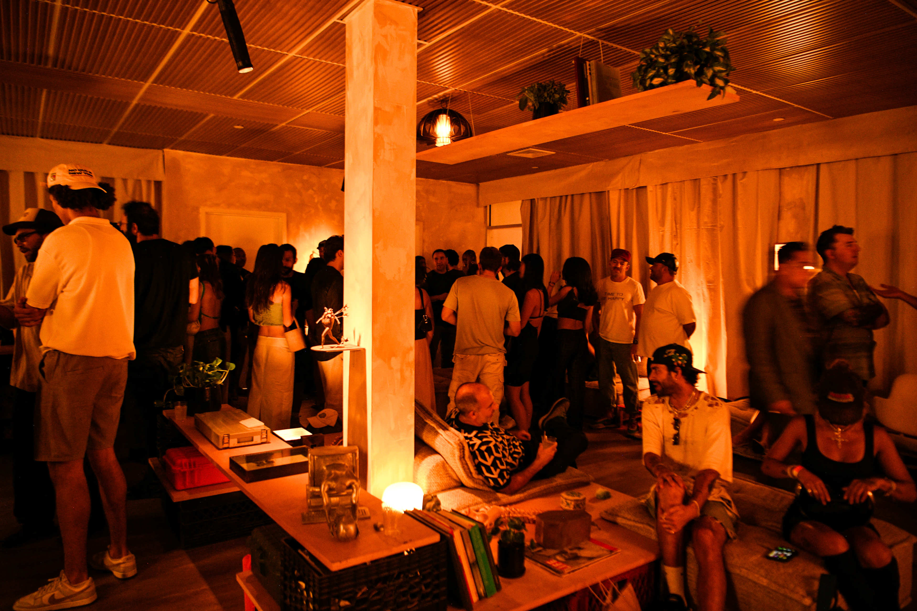 A crowded indoor space features people socializing amidst low lighting and various furniture arrangements.
