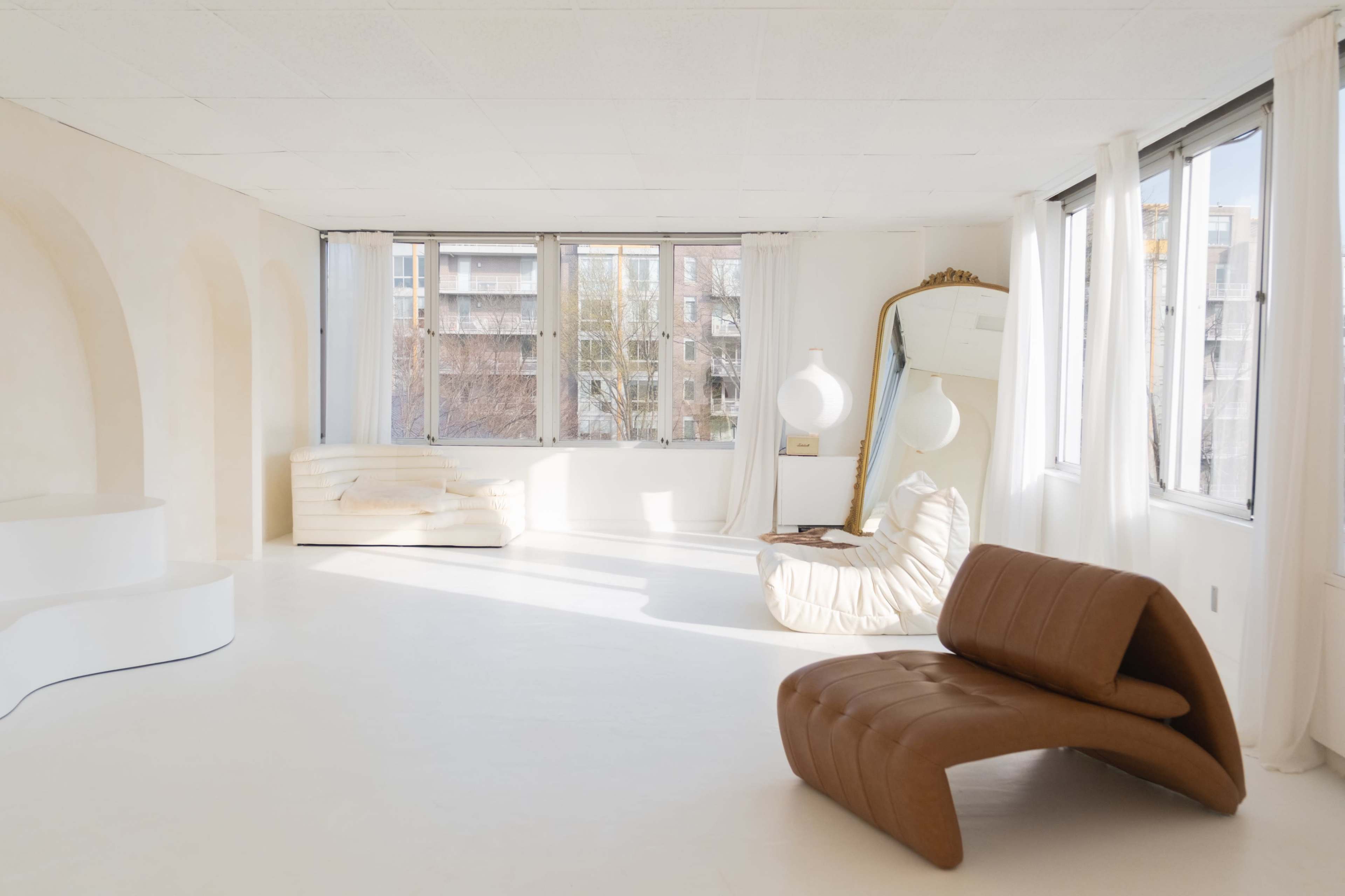 The image shows a spacious, minimalistic room with large windows, a brown curved sofa, a white chaise lounge, and a mirror.