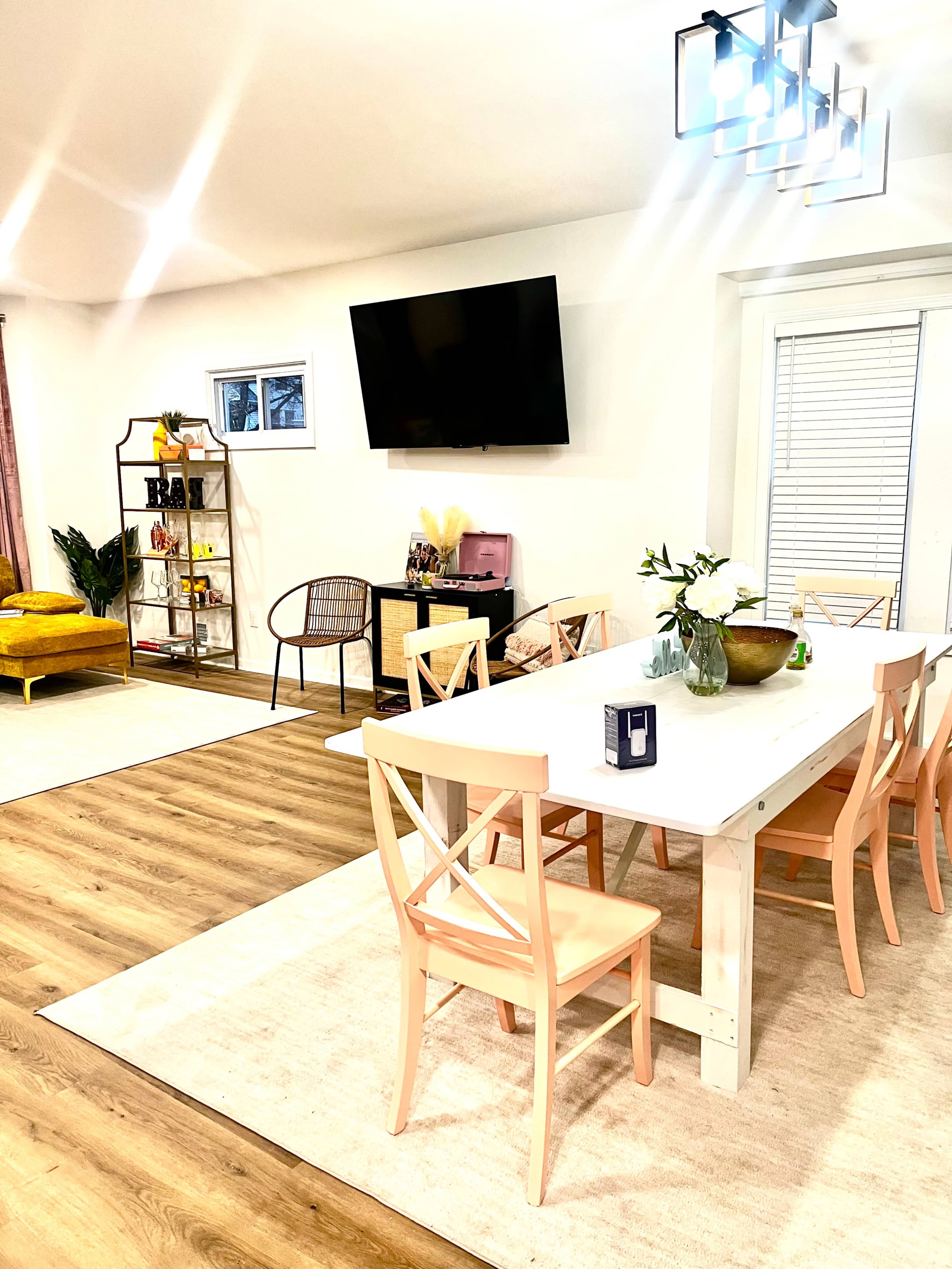A bright, modern living space featuring a dining table with pale chairs, a television mounted on the wall, and a seating area with a yellow couch.
