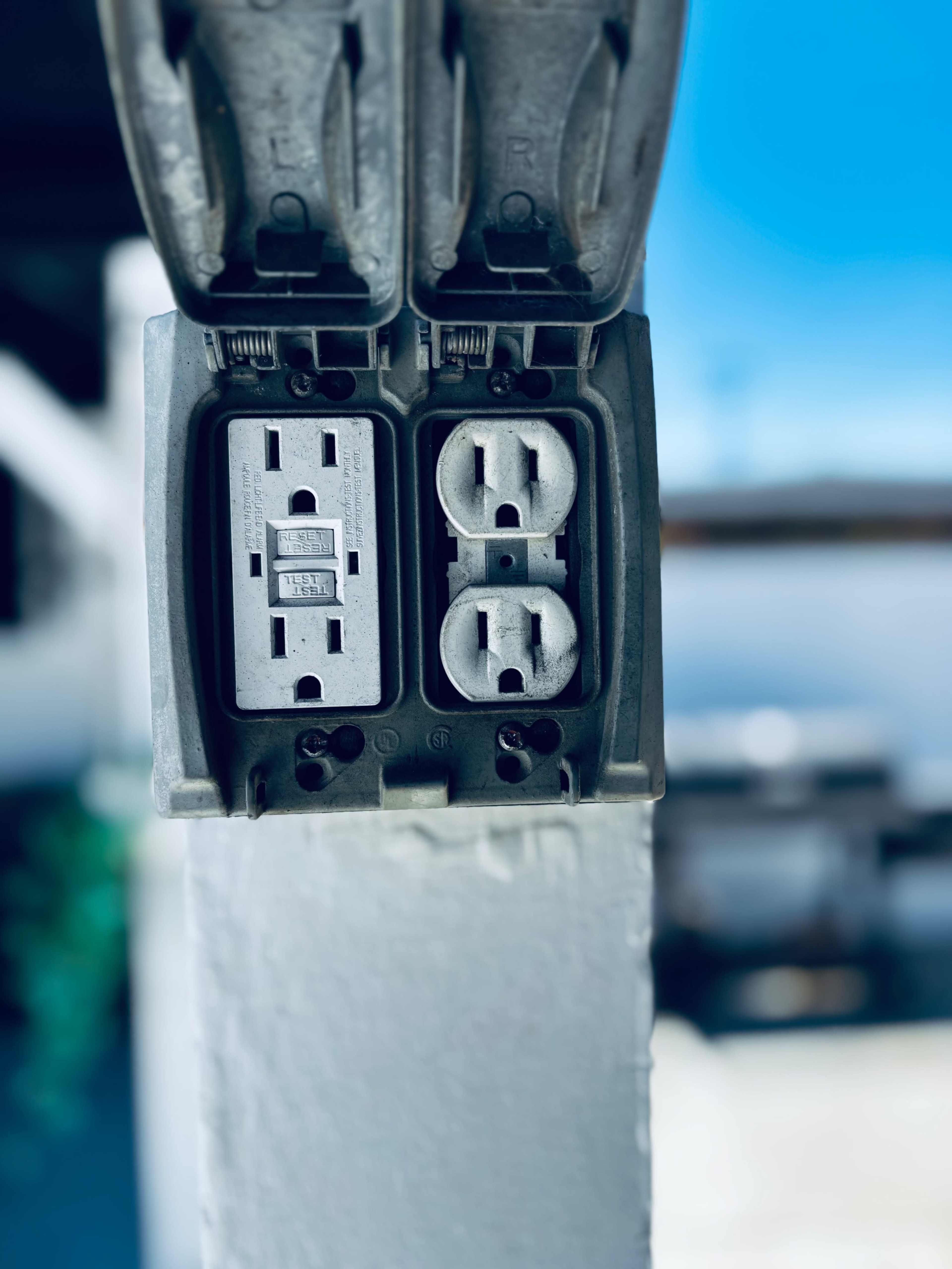 The image shows a weatherproof electrical outlet mounted on a post with two sockets visible.