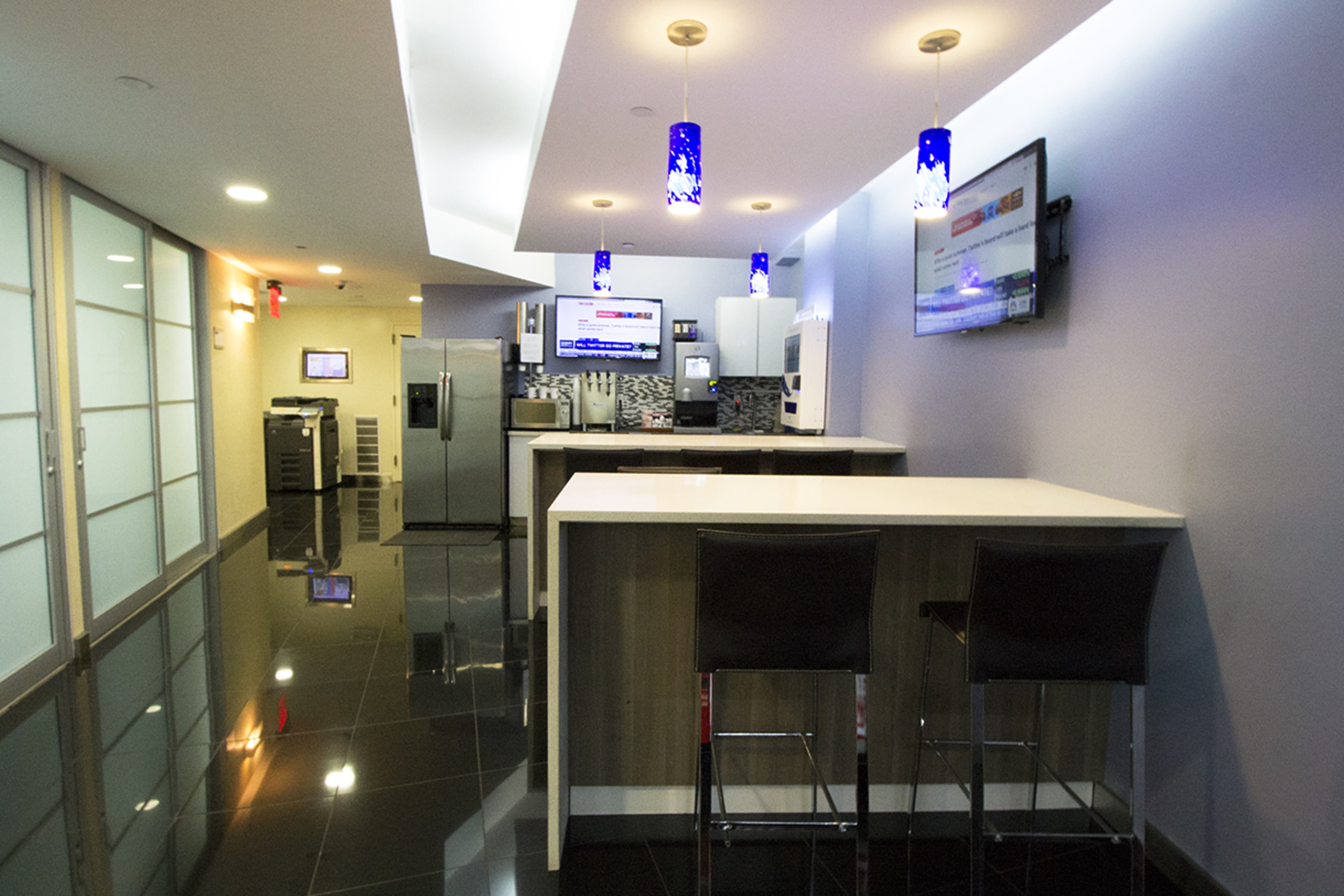 A modern kitchen area with a bar counter, high stools, stainless steel appliances, and flat-screen TVs mounted on the walls.