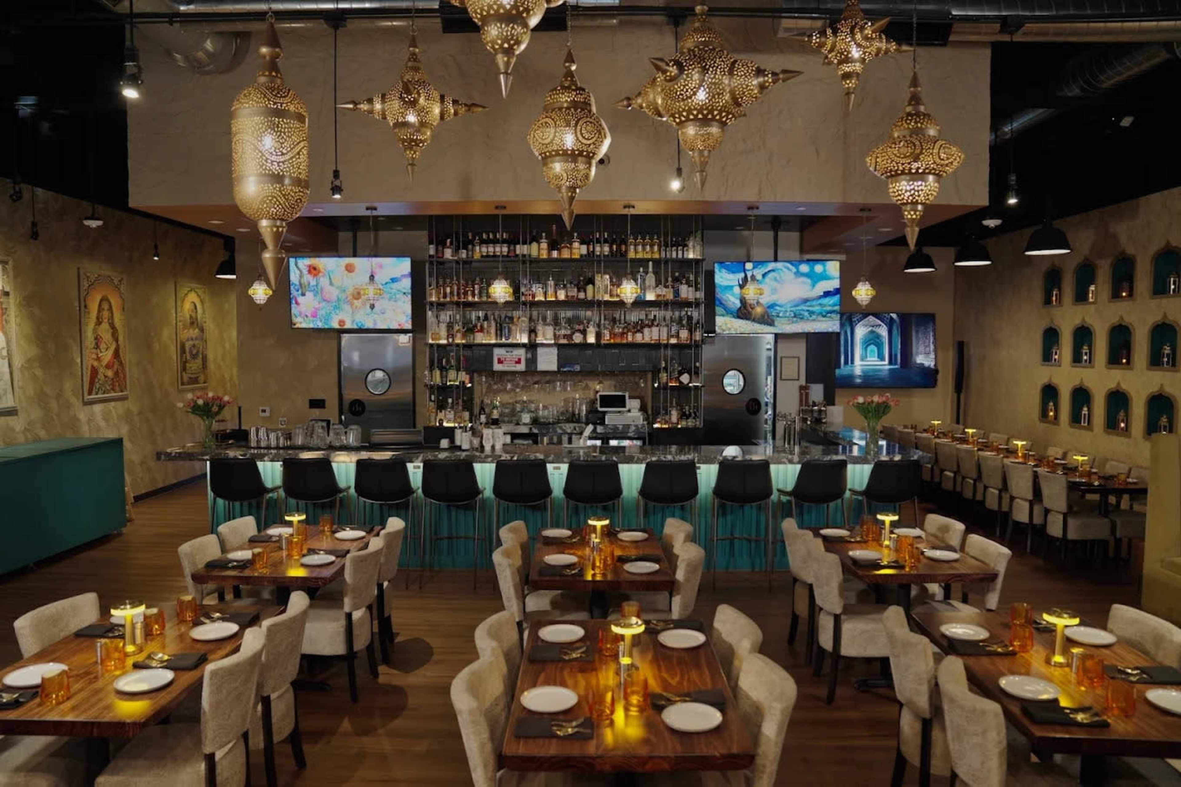 The image shows a well-decorated restaurant interior featuring a central bar area, elegantly arranged tables, and ornate hanging light fixtures.