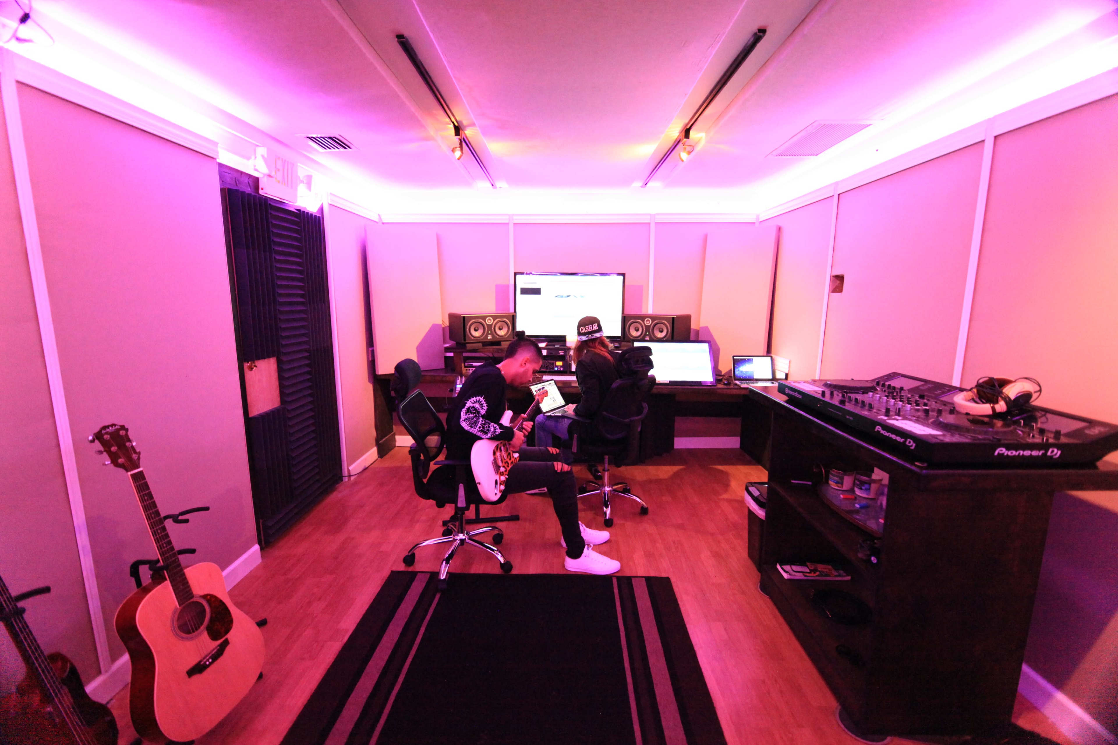 A musician plays guitar in a music studio with computers, speakers, and lighting enhanced by purple accents.