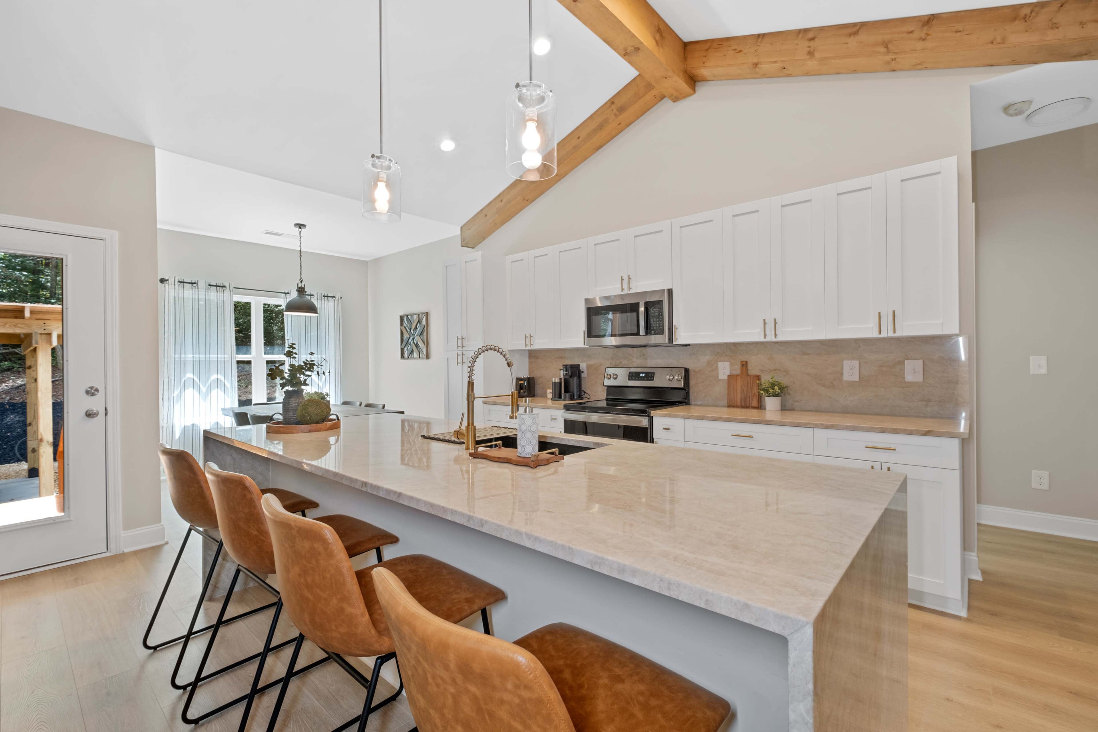 The modern kitchen features a central island with four brown bar stools, stainless steel appliances, and white cabinetry under wooden beams.