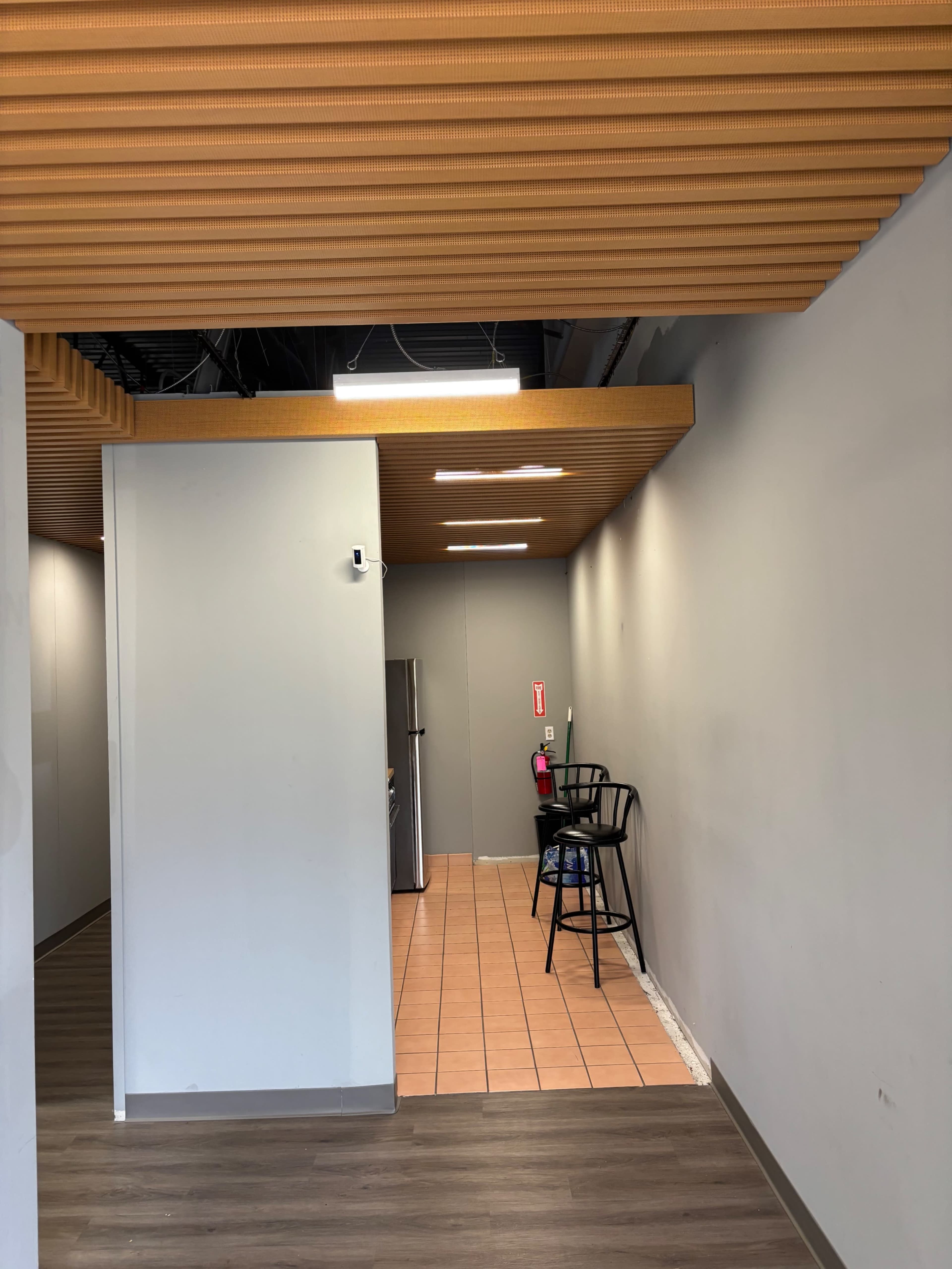 The image shows a partially finished interior space with a tiled floor, a kitchen area featuring a refrigerator, and a black chair positioned against the wall.